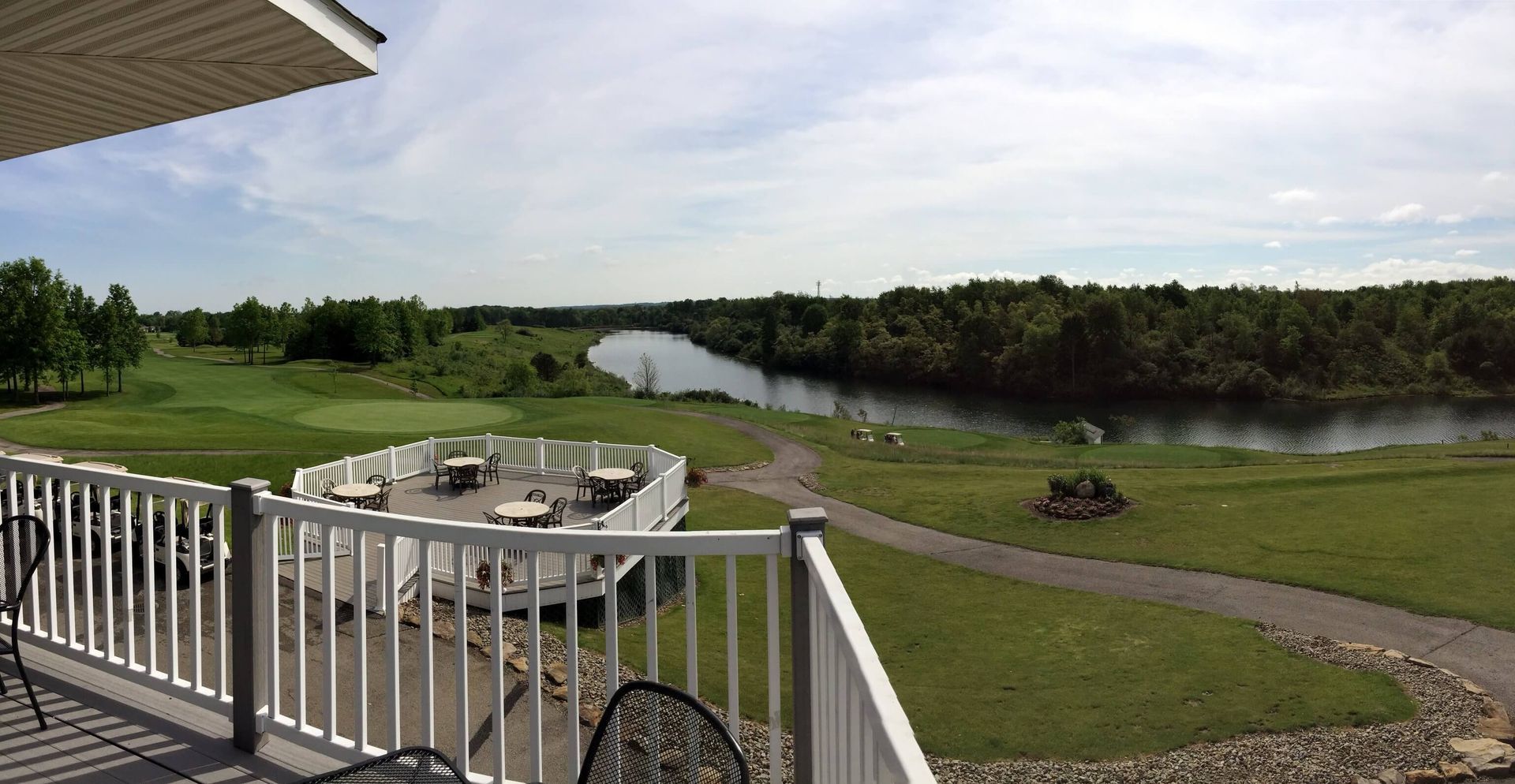 View of a golf course and river from a white deck with a railing. Green grass, trees, and cloudy sky.
