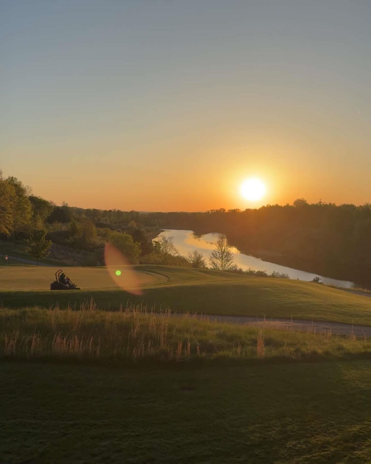 Sunset over a golf course. A lake reflects the golden light. A mower is on the green.