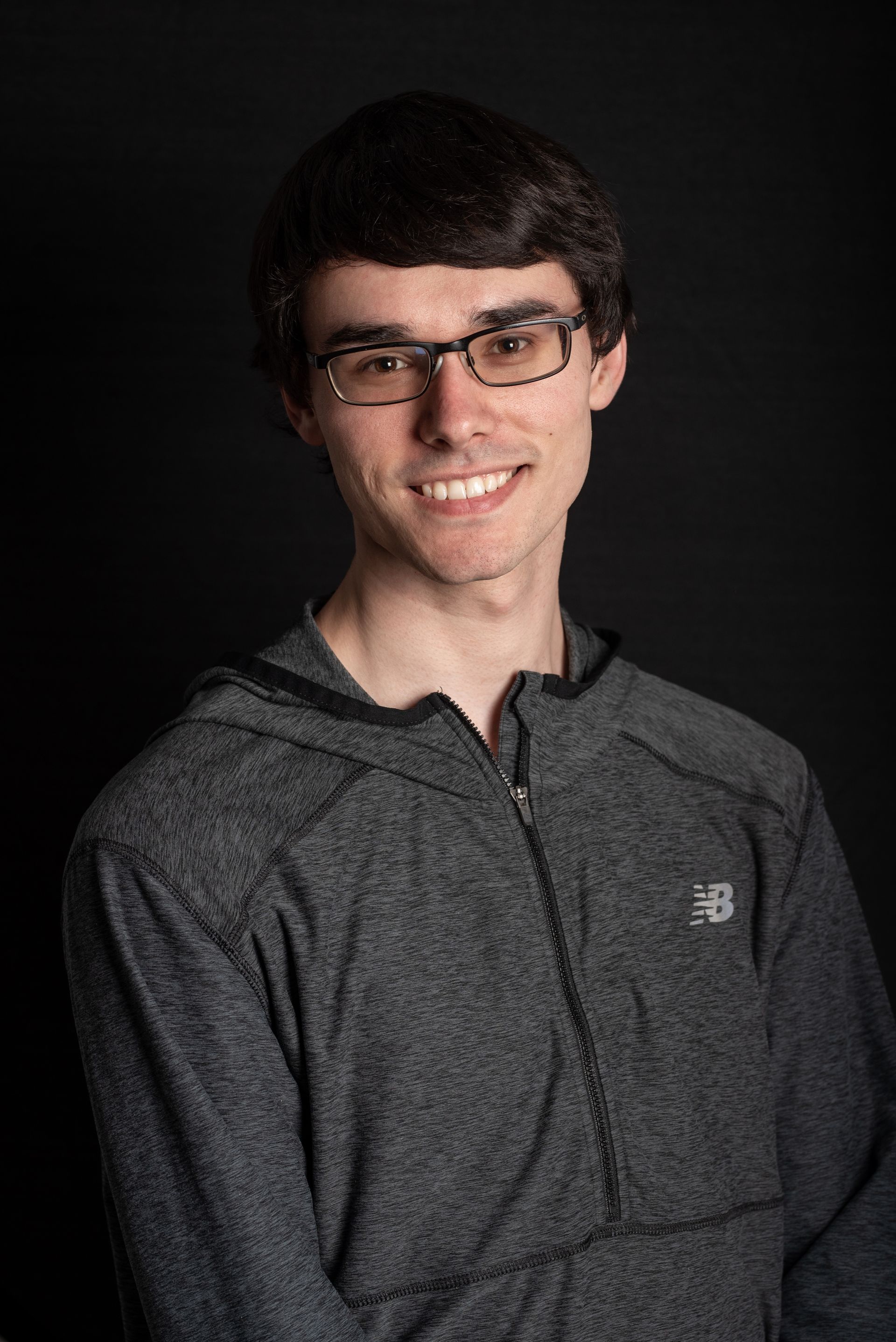 Un jeune homme portant des lunettes et un sweat-shirt à capuche gris sourit à la caméra sur un fond noir.