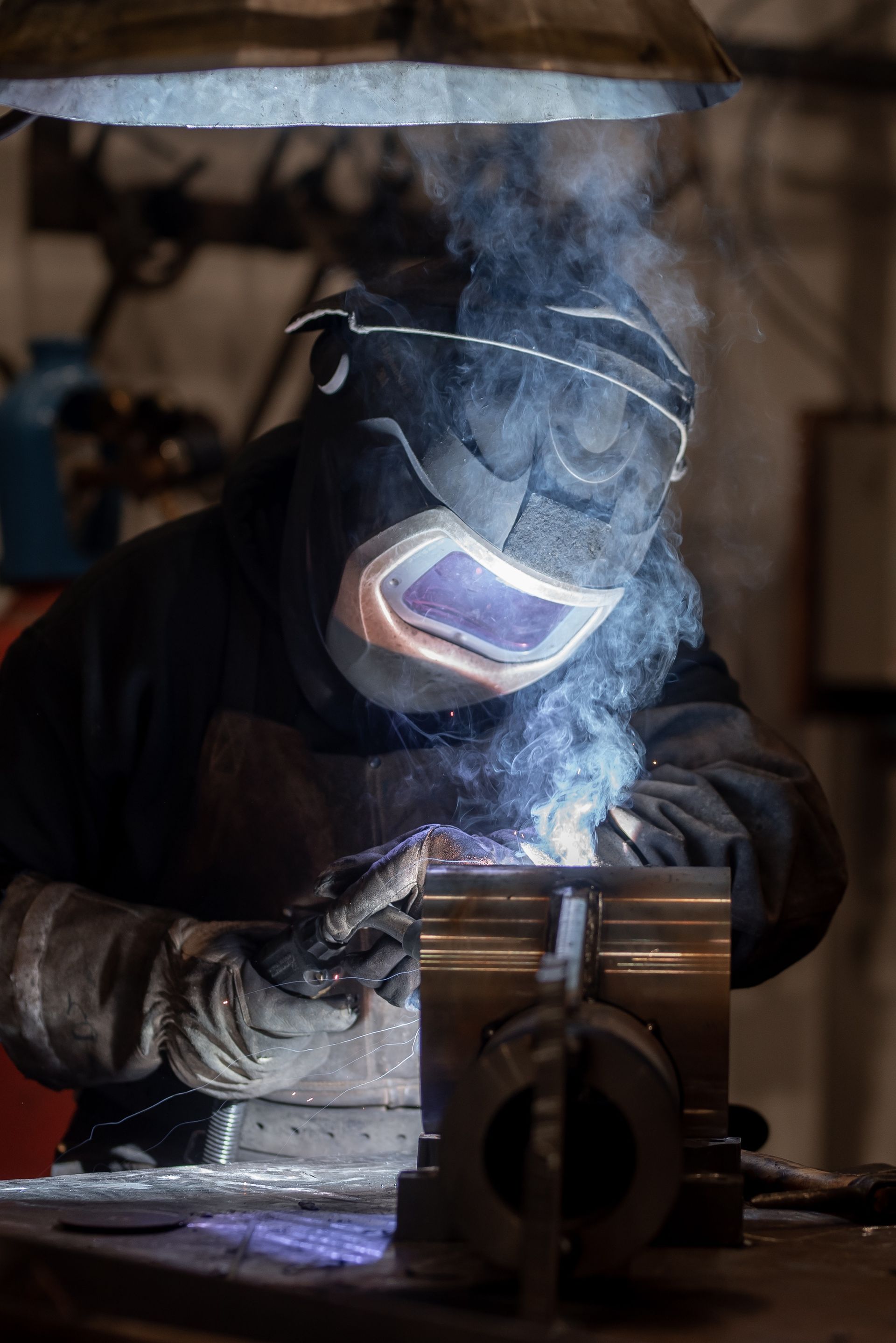 Welder in a dark workshop, wearing protective gear, welding metal, with sparks and smoke.