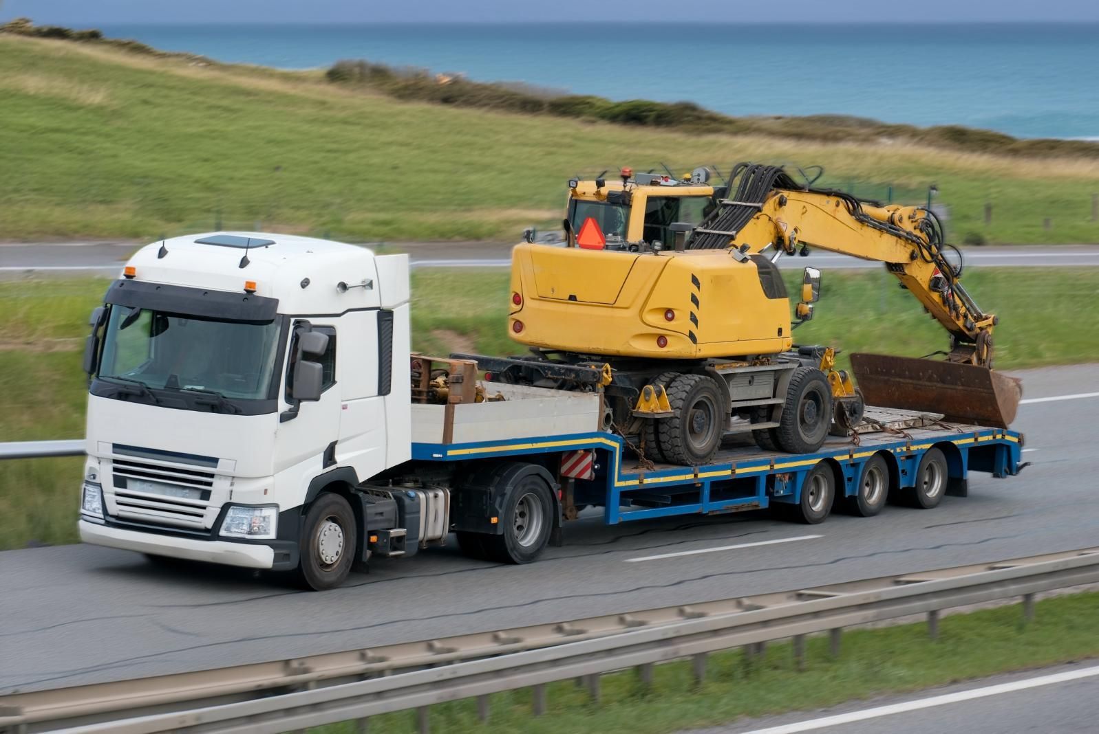 A Semi Truck With a Yellow Excavator on the Back is Driving Down a Highway — DJ's Tilt Tray Service in Coffs Harbour, NSW
