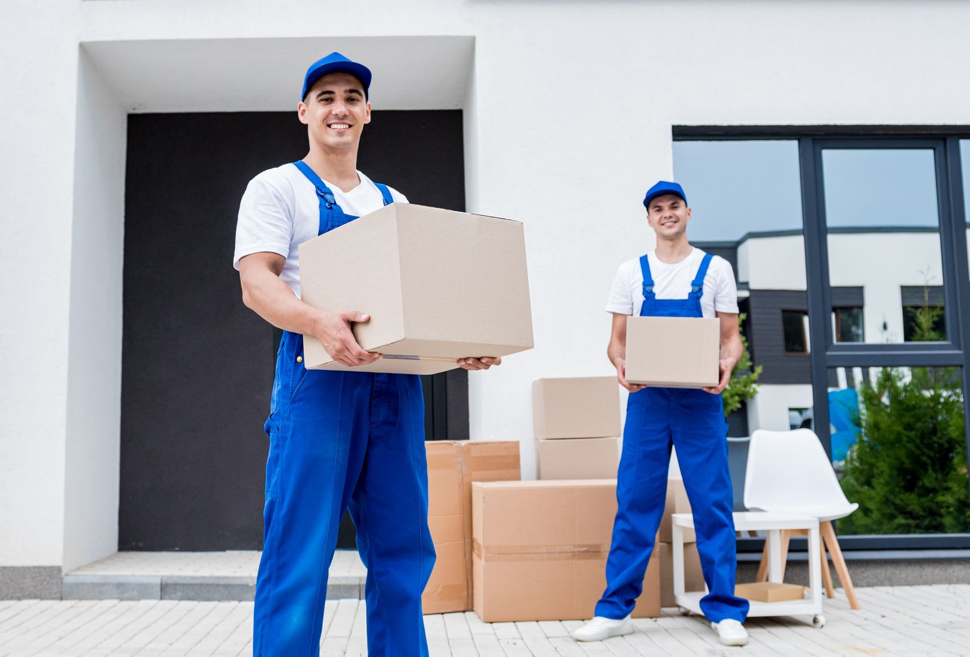 two men in blue overalls are holding cardboard boxes in front of a house .