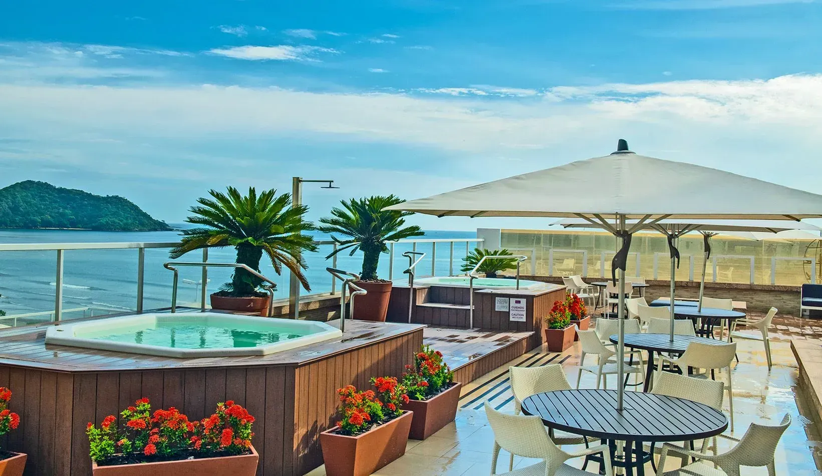 Terraço com jacuzzis, mesas e cadeiras com vista para o oceano sob um céu parcialmente nublado.
