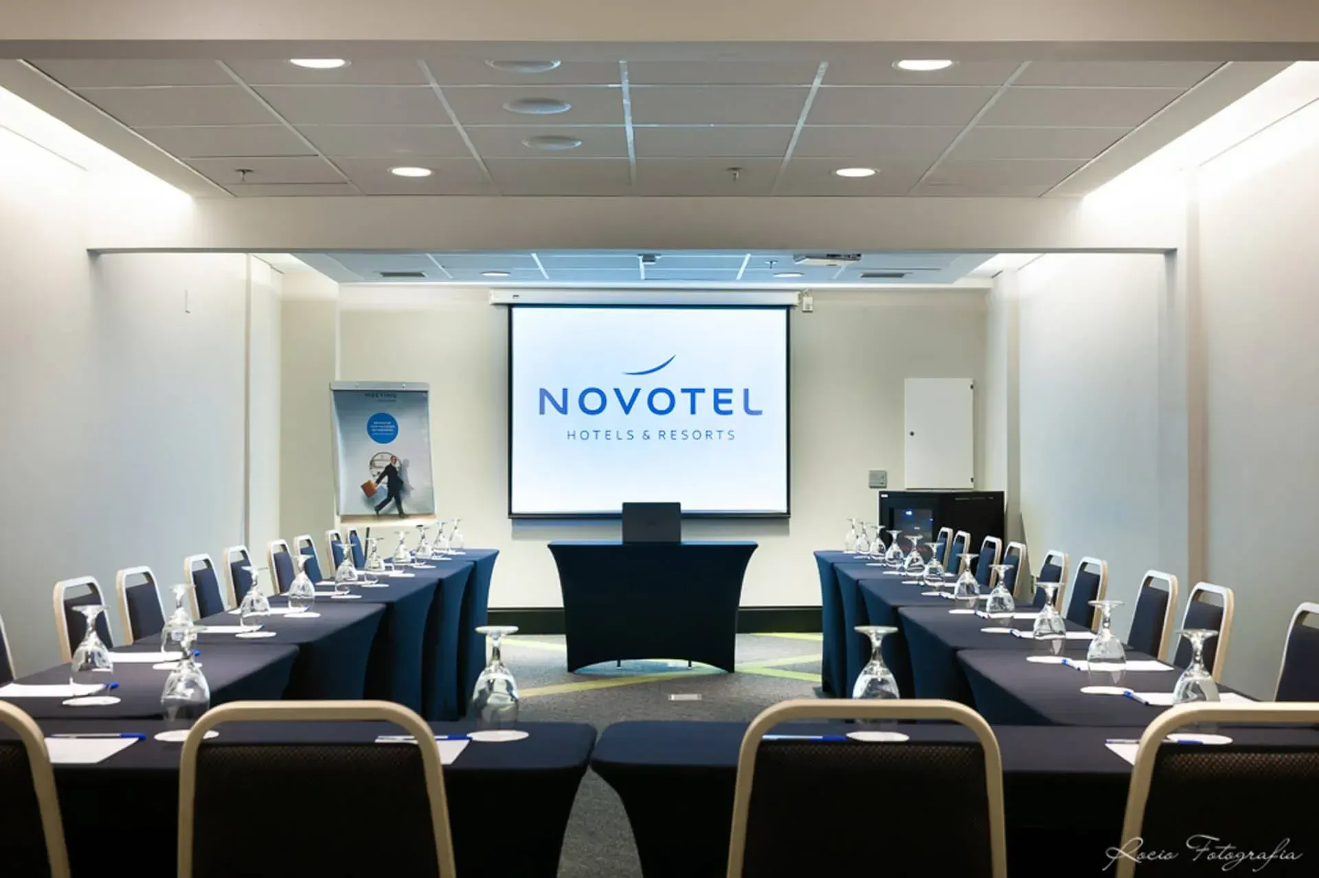 Uma sala de conferências com mesas e cadeiras e uma tela de projetor que diz novotel