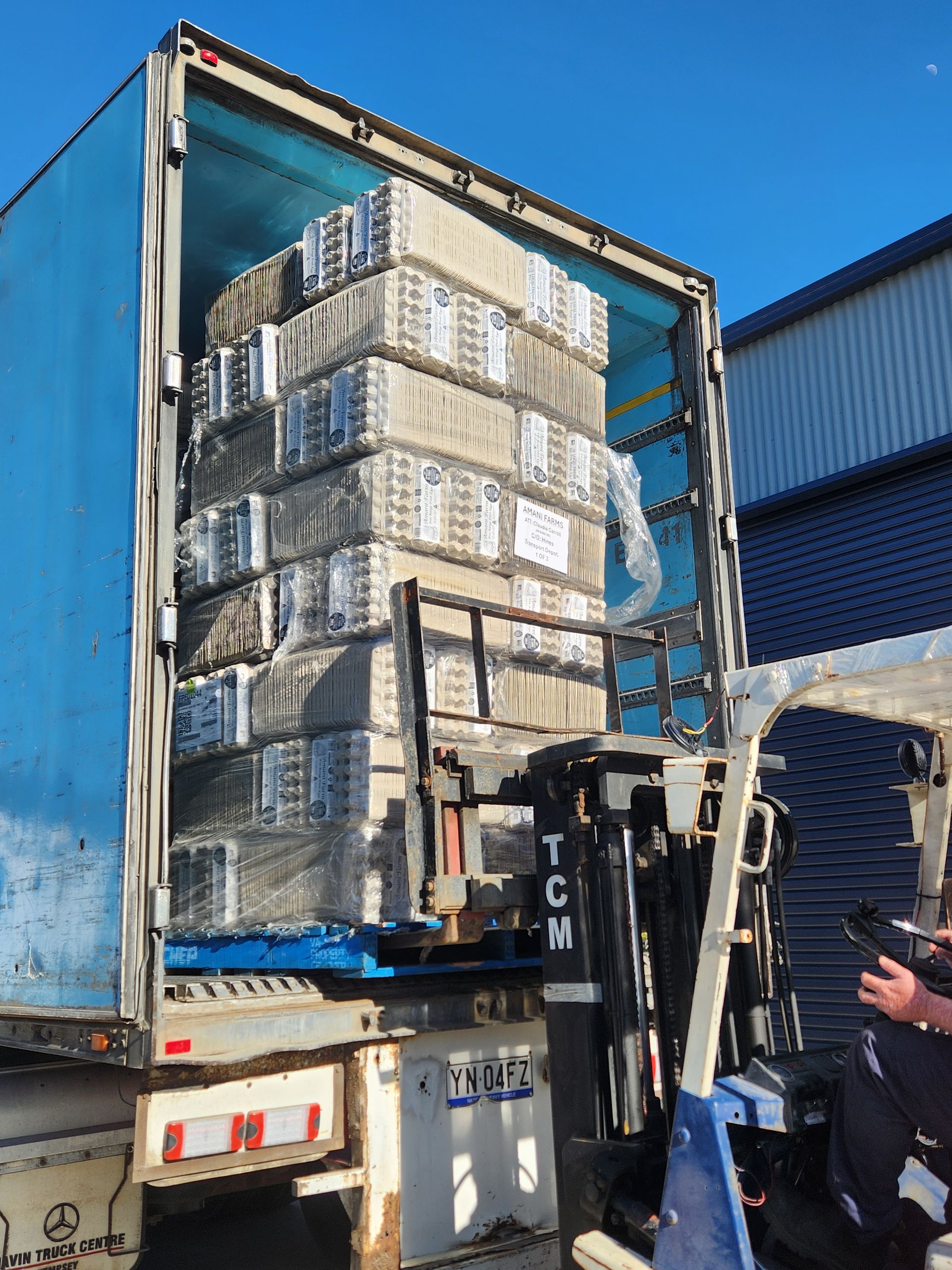 A forklift loading a tall, plastic-wrapped pallet of rectangular packages into the open trailer of a large truck.