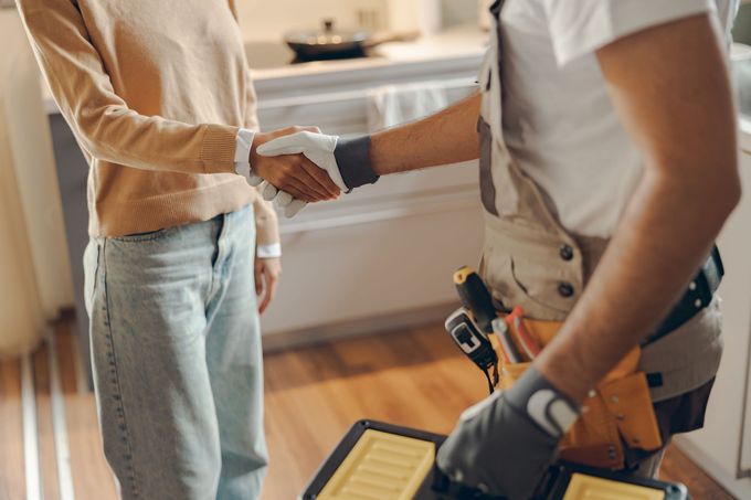 A person in a beige sweater shakes hands with a service worker wearing gloves and a tool belt in a residential kitchen.