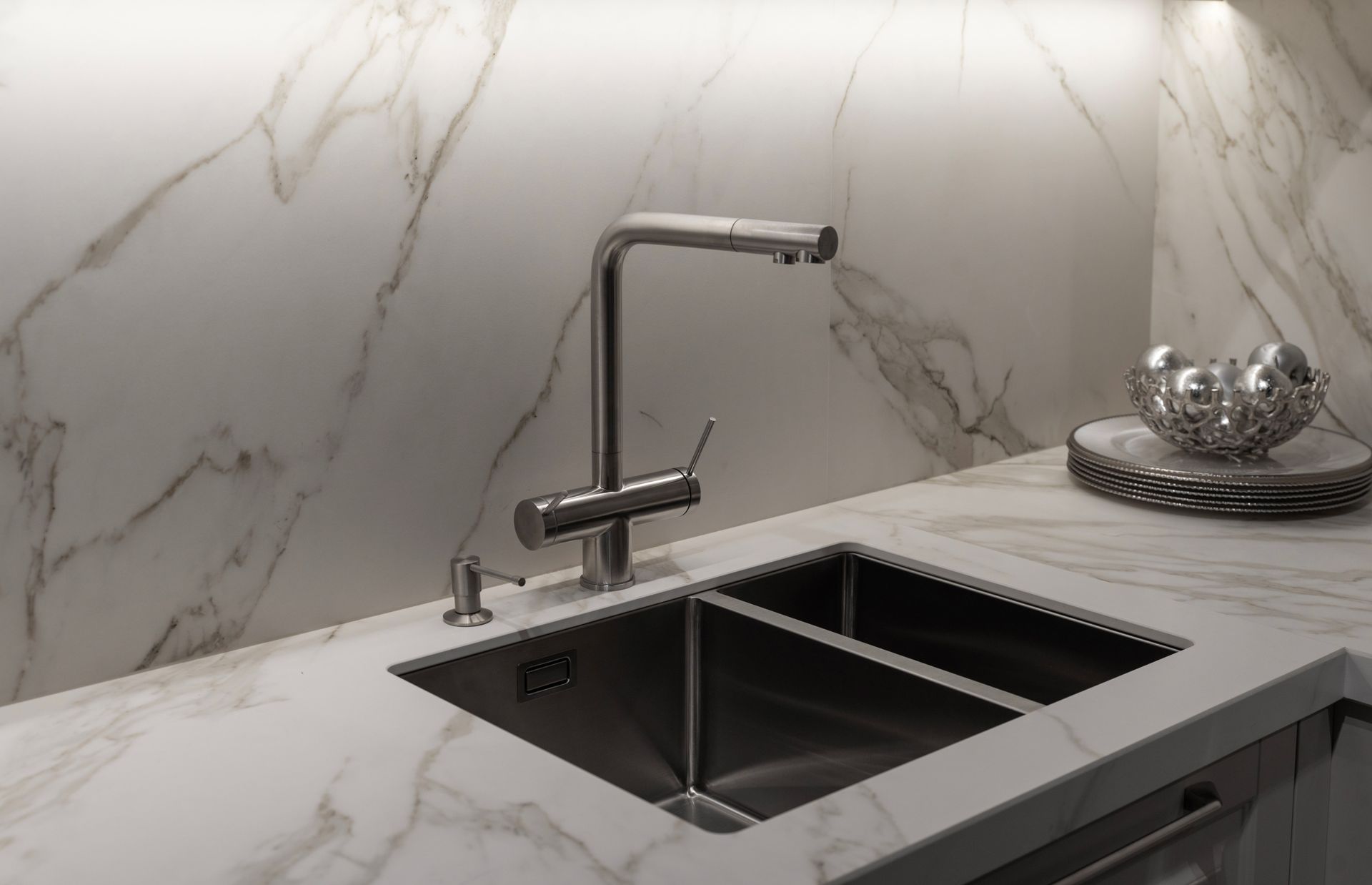 A stainless steel faucet and dual-basin sink mounted in a white marble countertop with a matching marble backsplash.