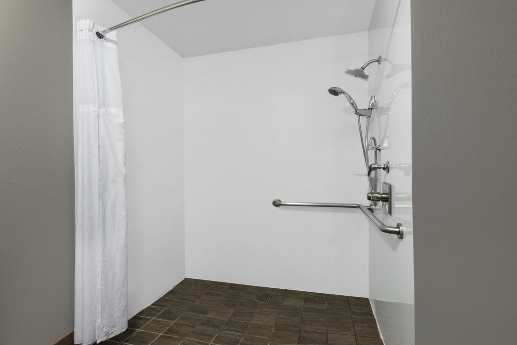 Accessible shower stall with grab bar, shower head, and curtain; brown tiled floor, white walls.