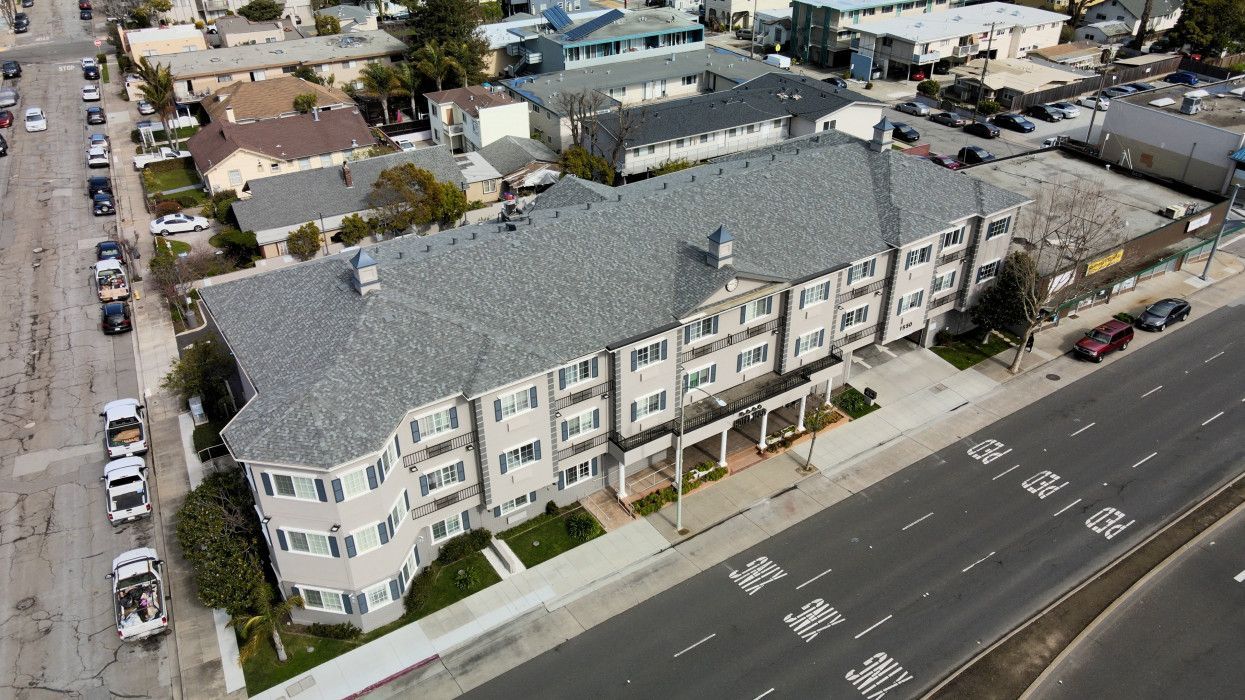 Aerial view of a three-story beige apartment building with a grey roof next to a multilane road.