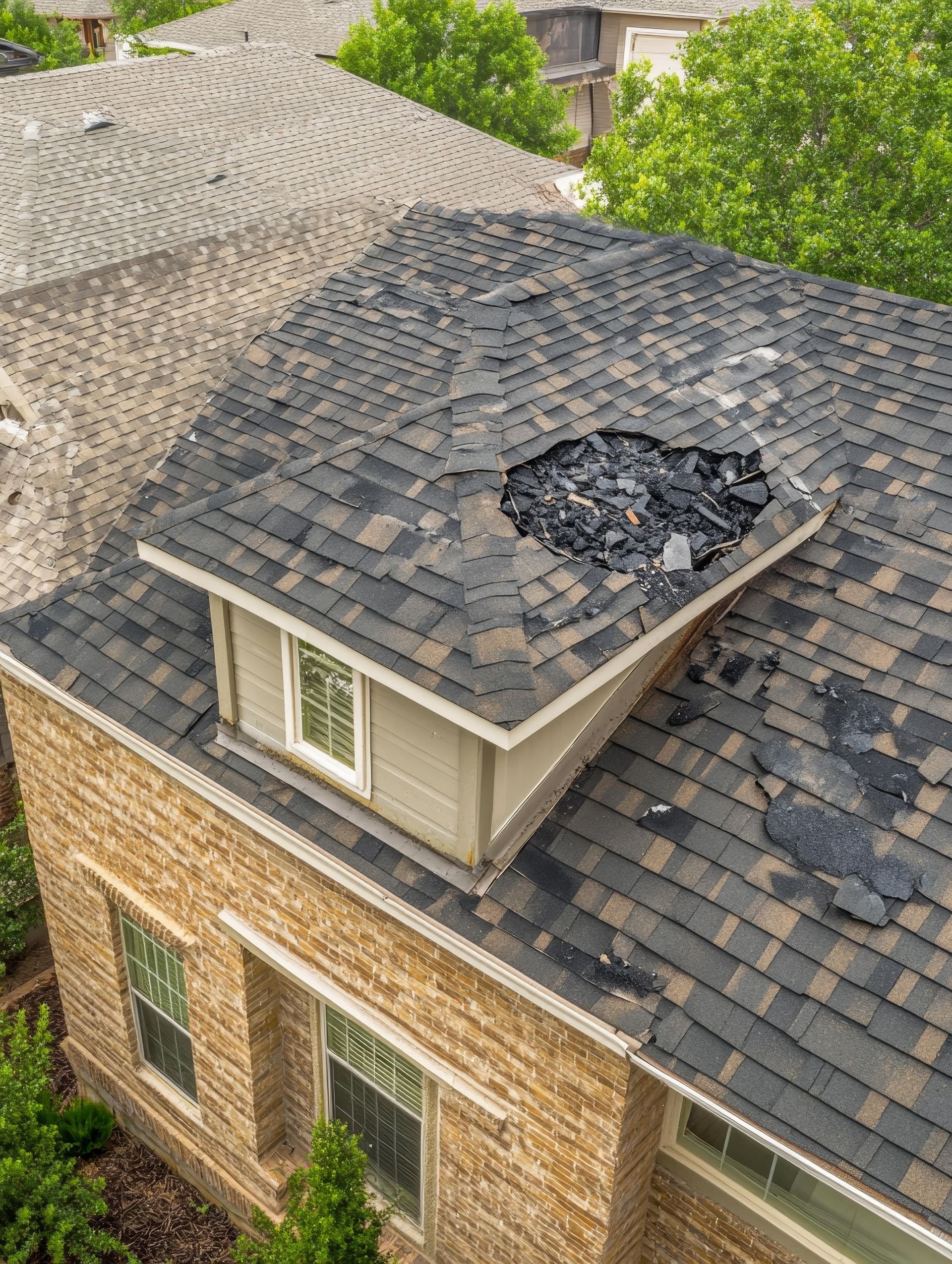 Damaged residential roof with a large hole. Brown and black shingles, tan dormer.