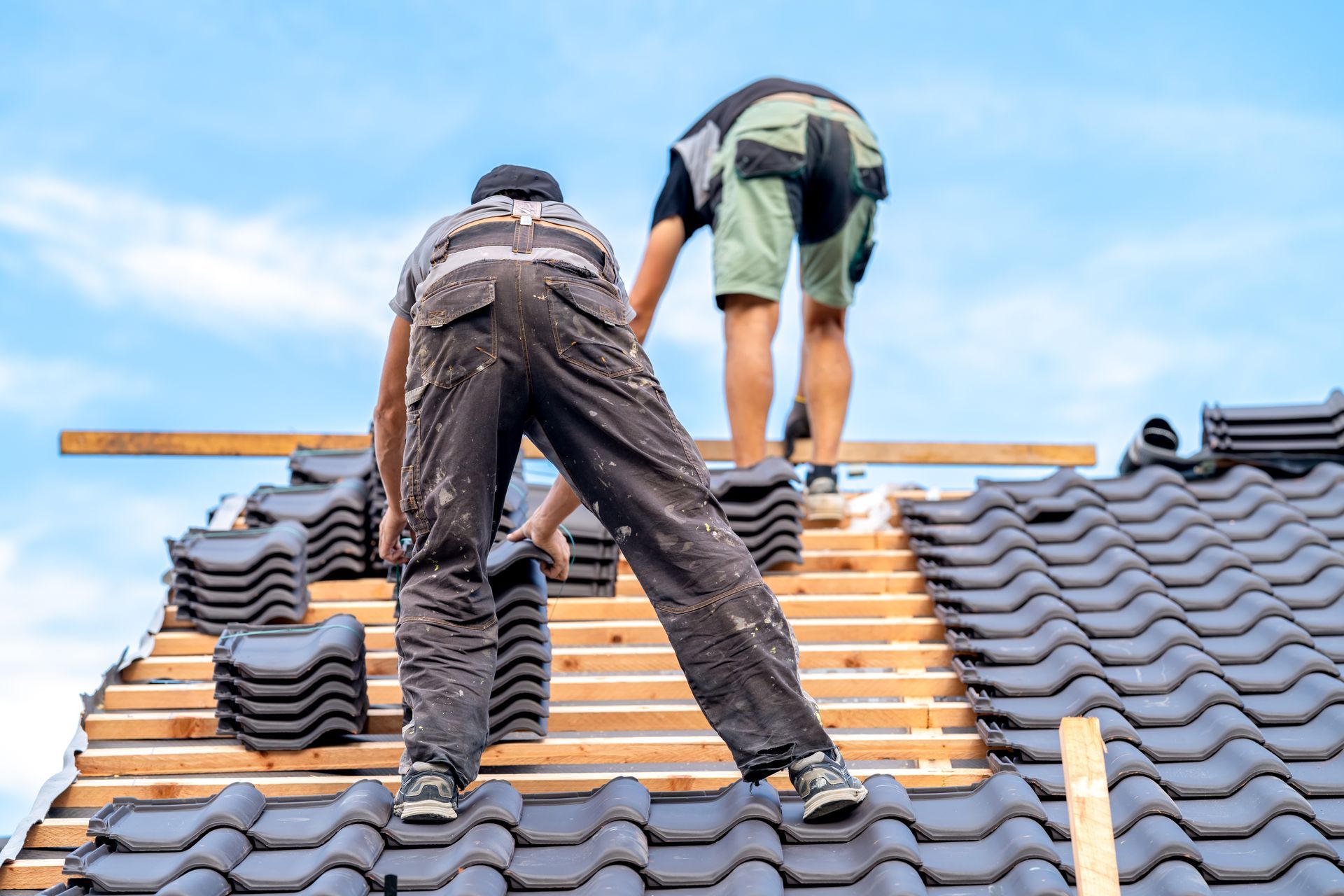 Two roofers installing black tiles on a roof against a blue sky.