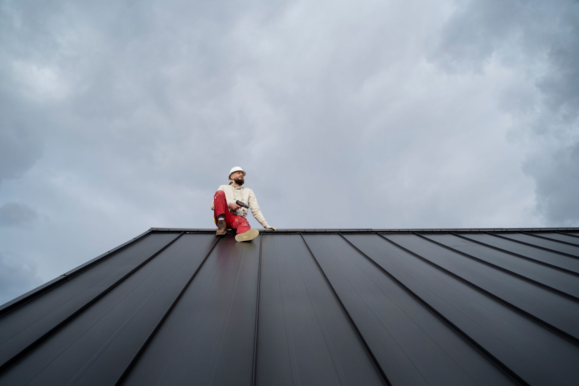 Roofer on a dark metal roof under a cloudy sky. Man in safety gear.
