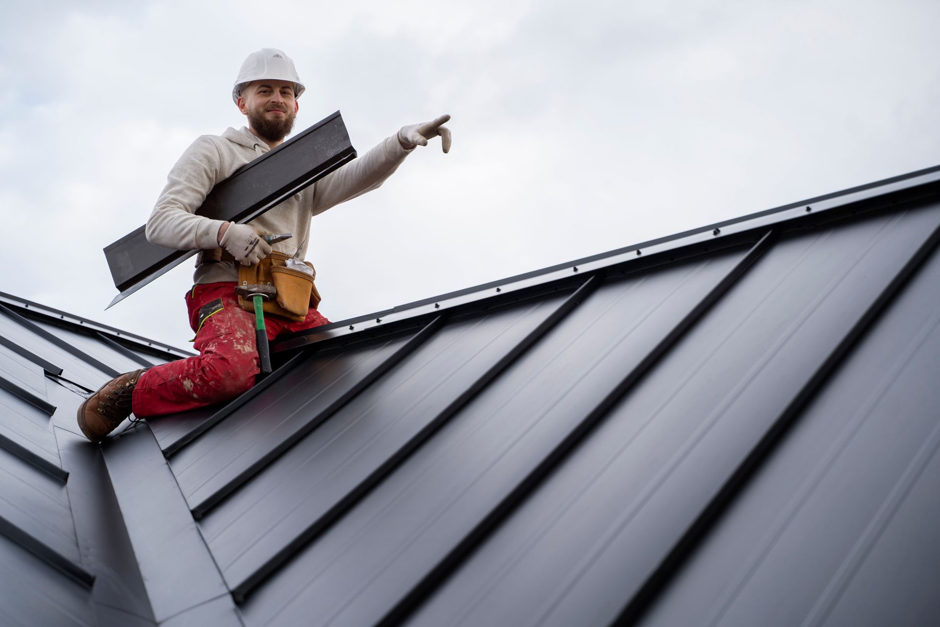 Roofer on a dark metal roof, pointing; holds a piece of material, wears a hard hat and tool belt.