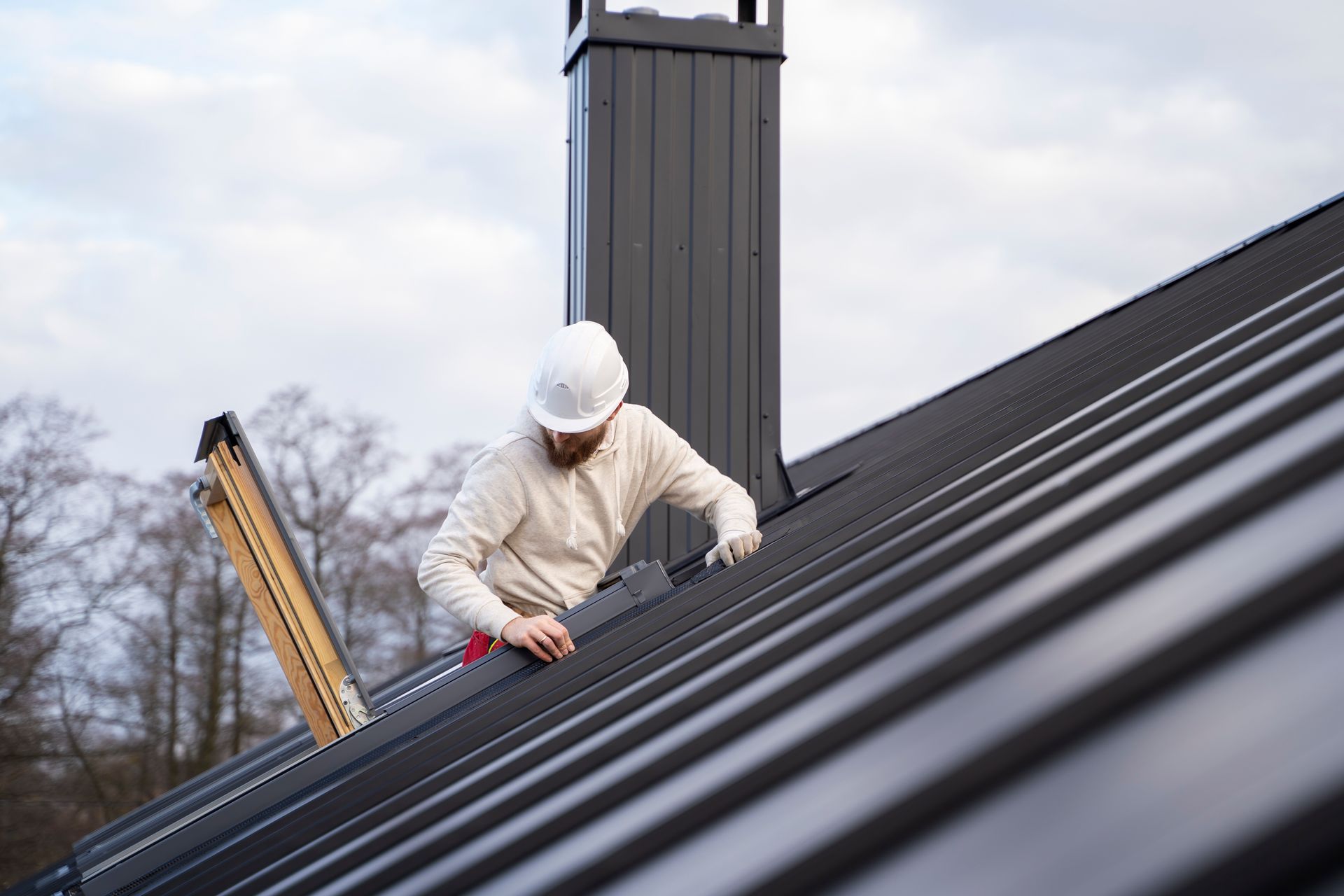 Roofer on a dark metal roof, working near a chimney, wearing a hard hat, ladder nearby, overcast sky.