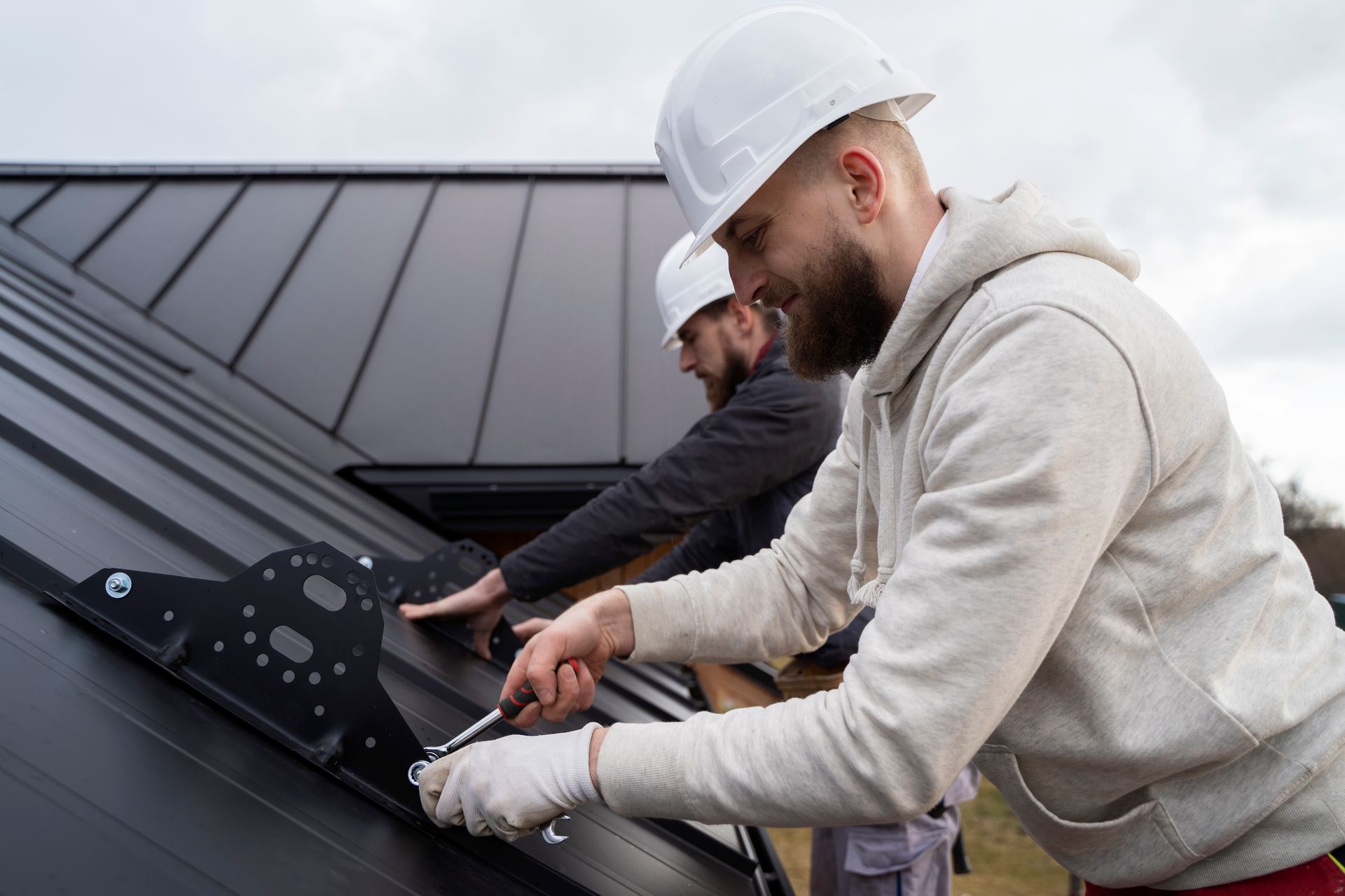 Two roofers in hard hats installing a black metal roof on a cloudy day.