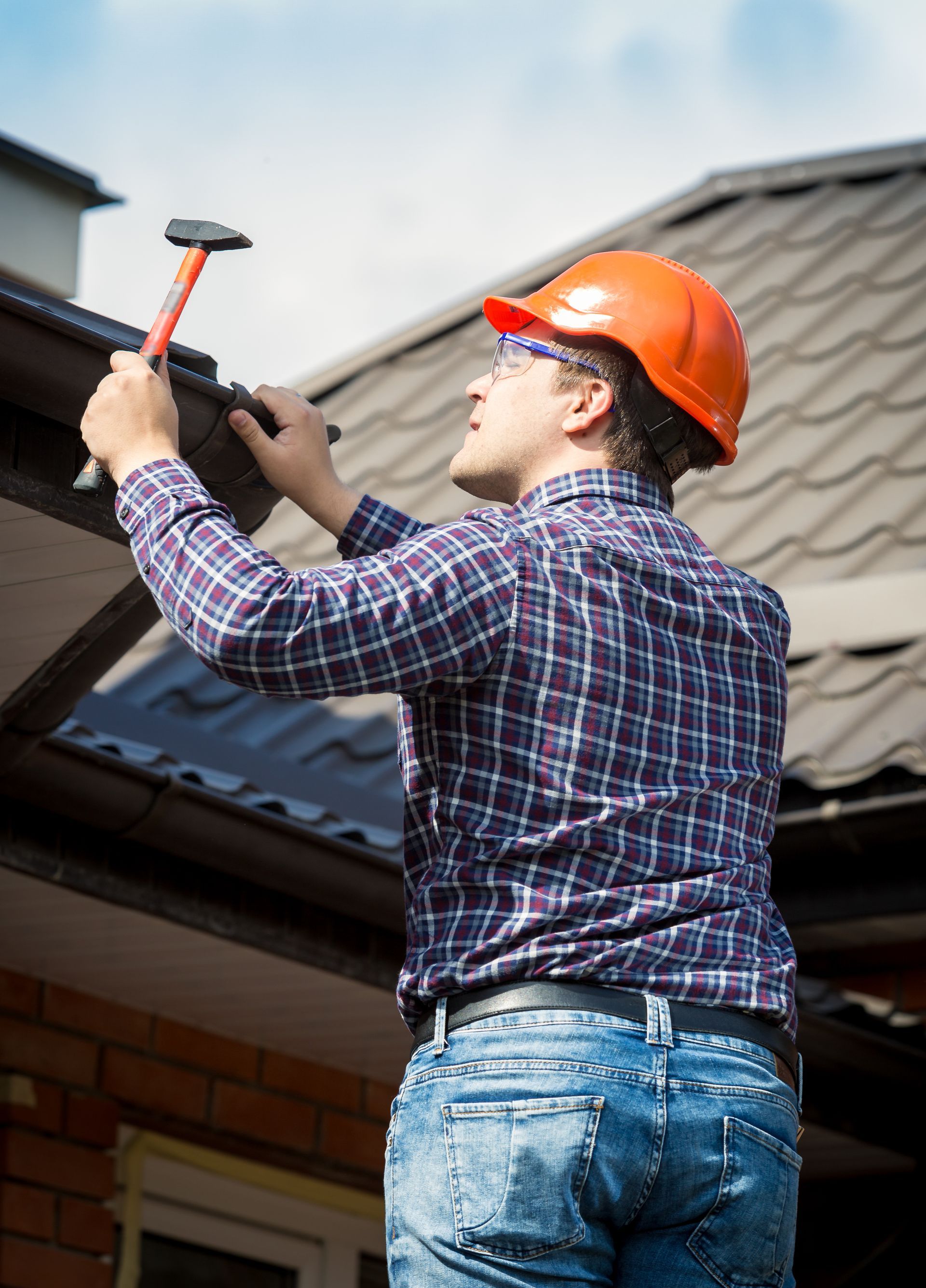 Man in orange hard hat hammers on a roof gutter.
