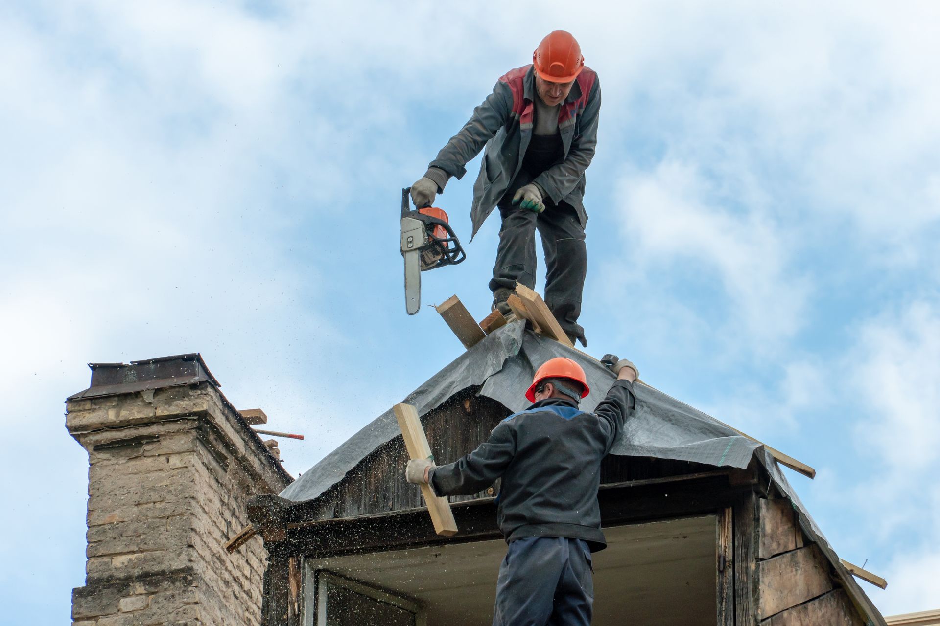 Two construction workers on a roof, one using a chainsaw, the other holding a board; blue sky background.