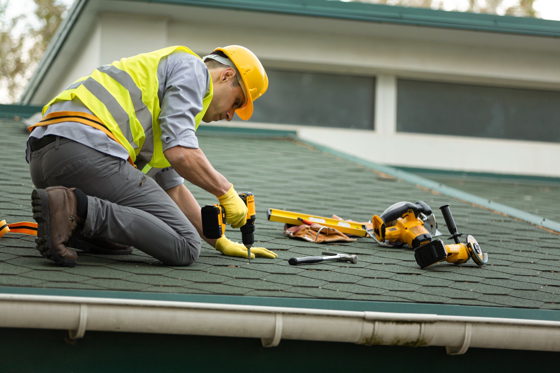 Roofer on a green roof, wearing a yellow hard hat and vest, using a power drill next to tools.