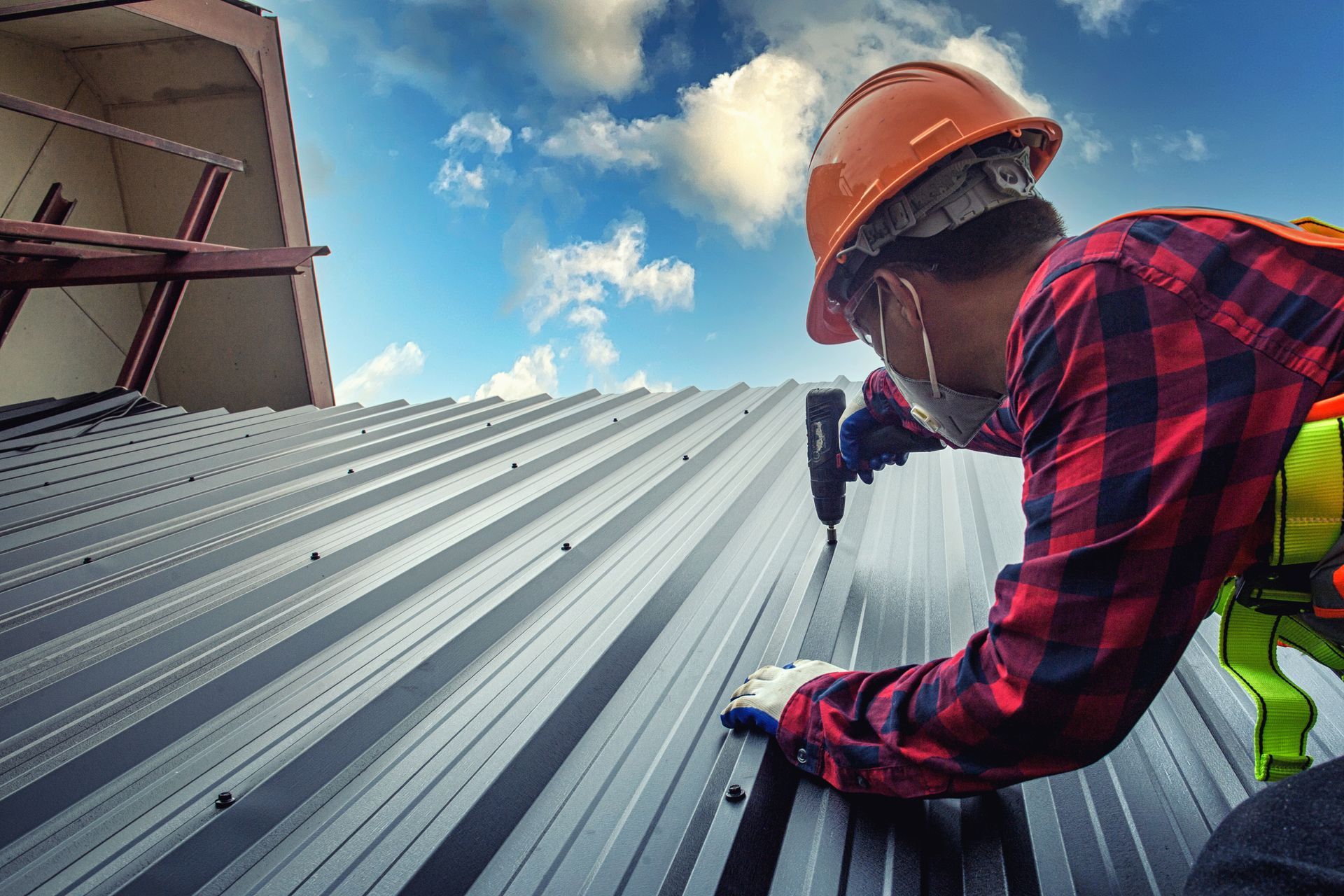 Construction worker in hard hat, installing a metal roof panel on a building under a cloudy blue sky.