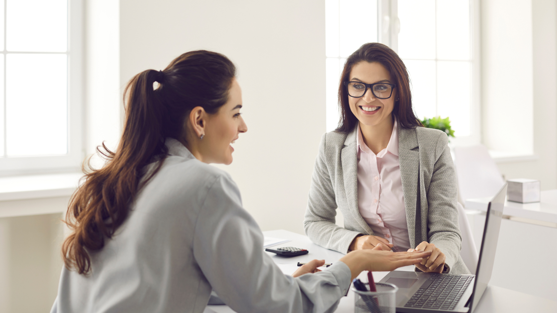 Two women are sitting at a table with a laptop and talking to each other.