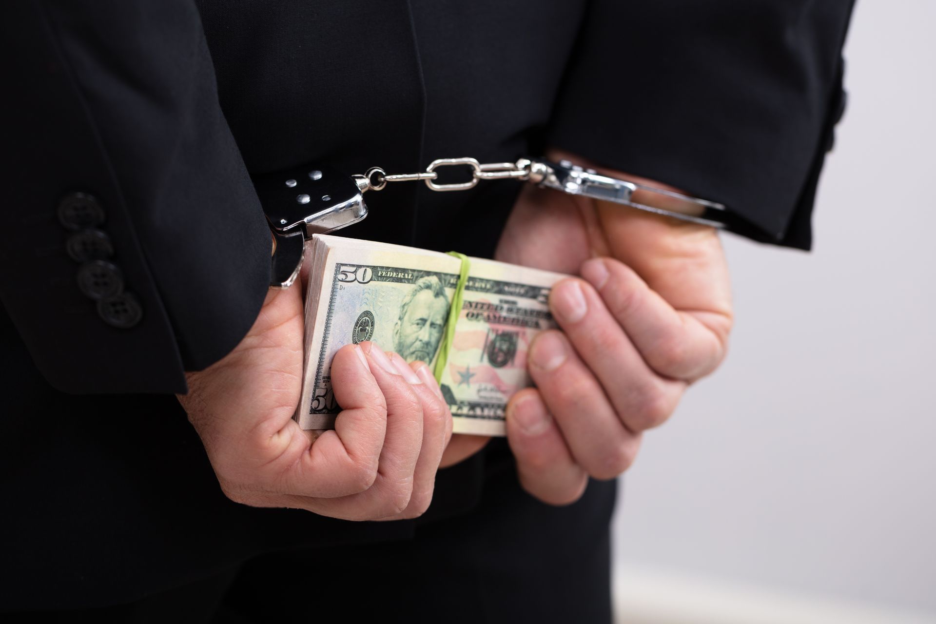 Person in handcuffs holding a stack of fifty-dollar bills behind their back.