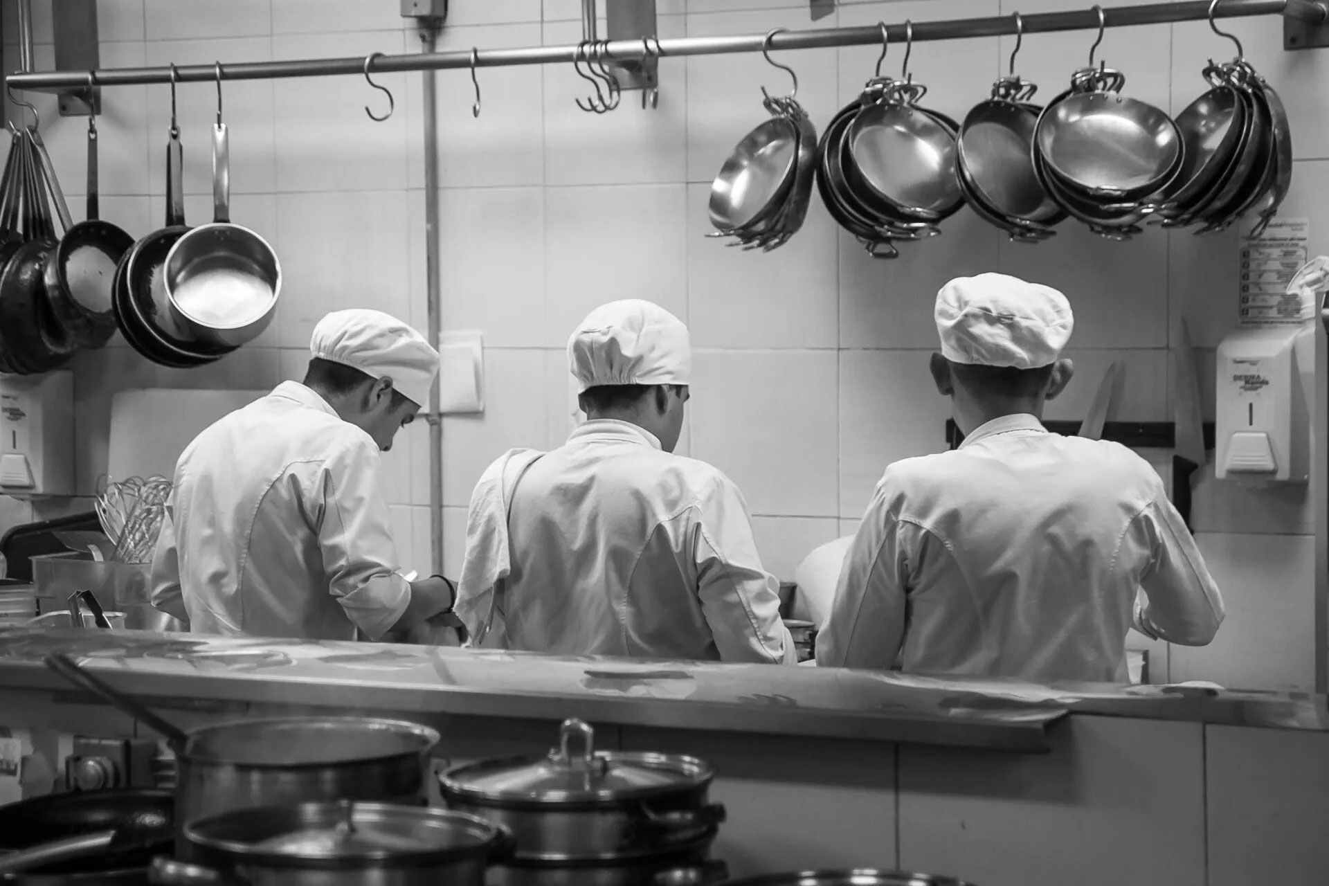 Tres chefs con uniformes blancos cocinando en una cocina industrial con ollas y sartenes colgadas.
