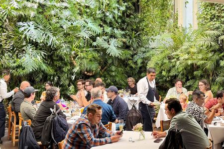 Personas sentadas en mesas redondas en una exuberante cafetería con jardín interior, con un camarero que les llevaba bebidas.