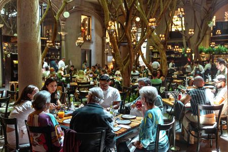 Patio de restaurante abarrotado con comensales sentados en mesas bajo cálidas luces colgantes y árboles.