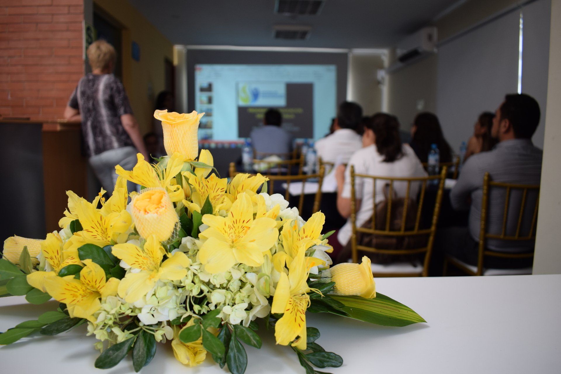 Un ramo de flores amarillas está sobre una mesa frente a un grupo de personas sentadas en mesas.