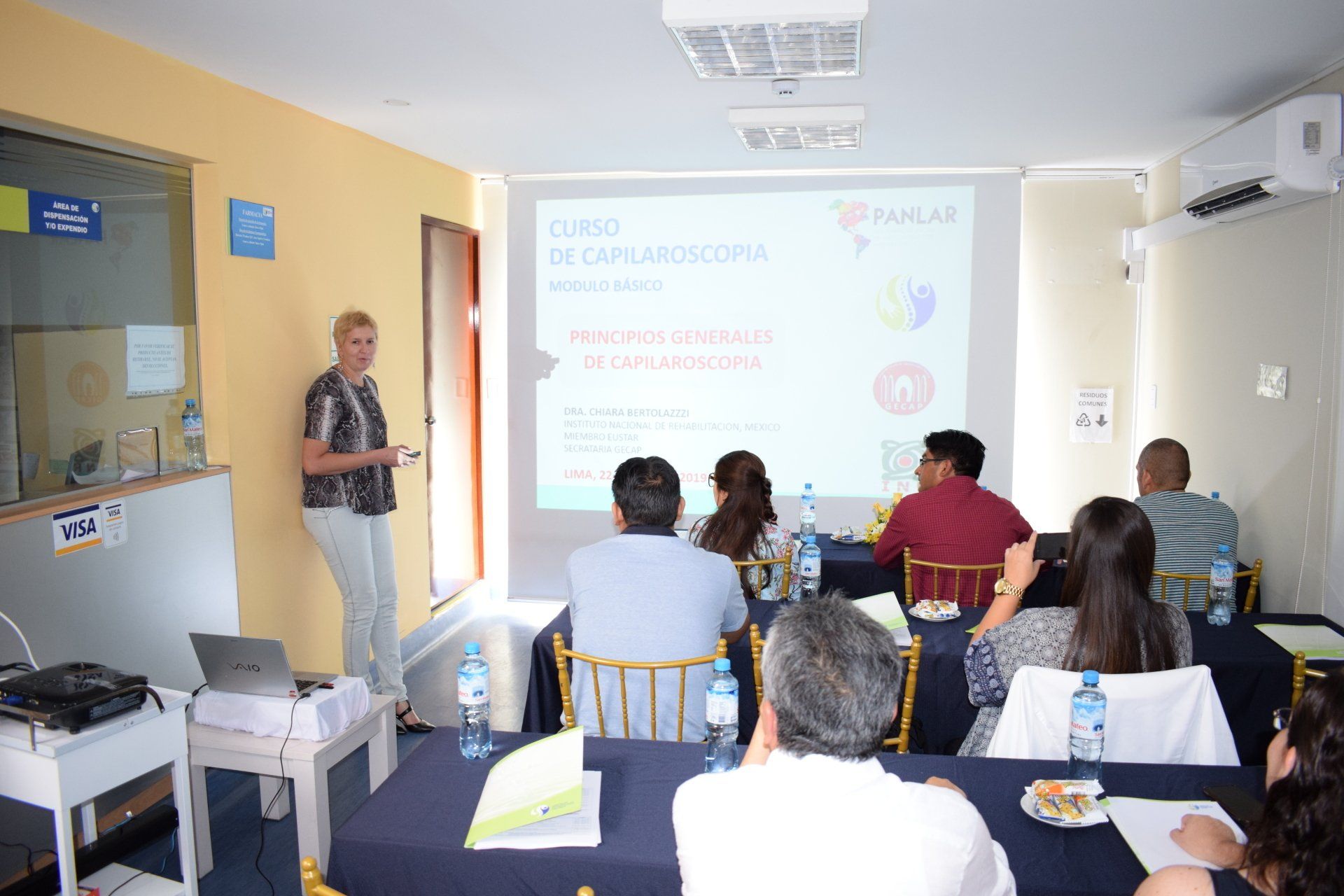 Una mujer está dando una presentación a un grupo de personas sentadas en mesas.