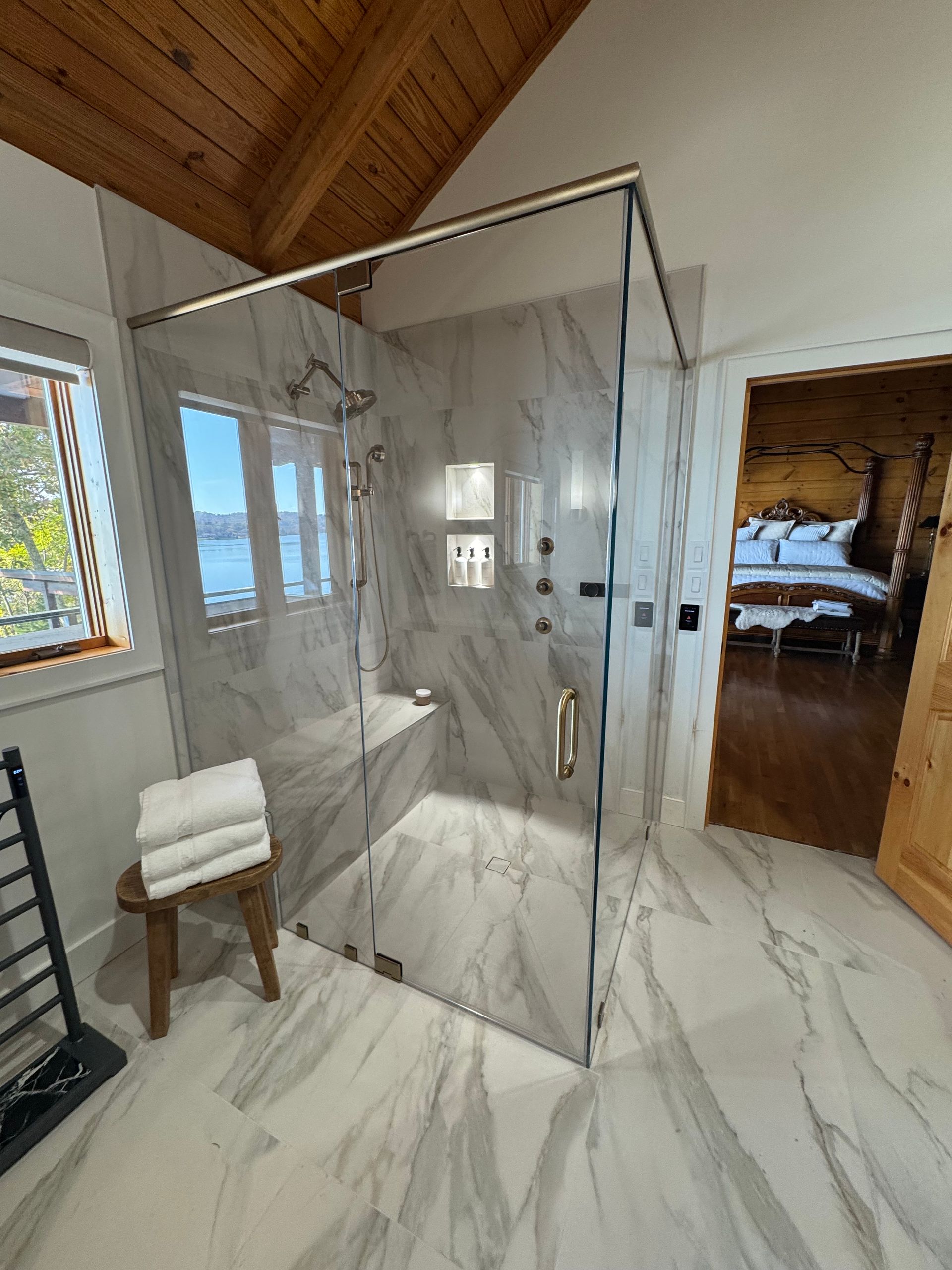 Spacious bathroom with marble tile and glass shower, wooden ceiling, view of water. Towels on stool, heated towel rack, 