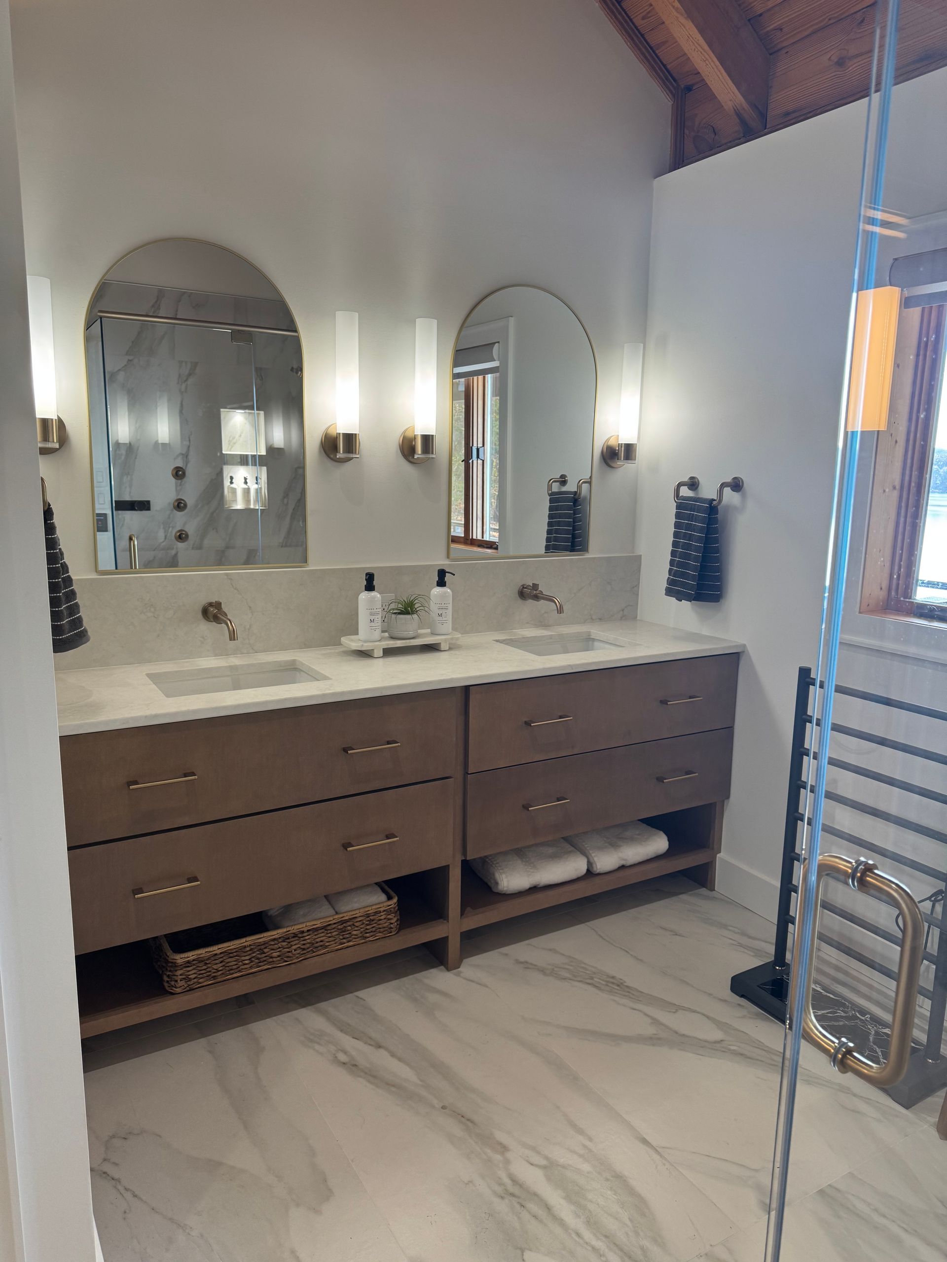 Bathroom with dual sinks, arched mirrors, gold sconces, and a marble-look floor.