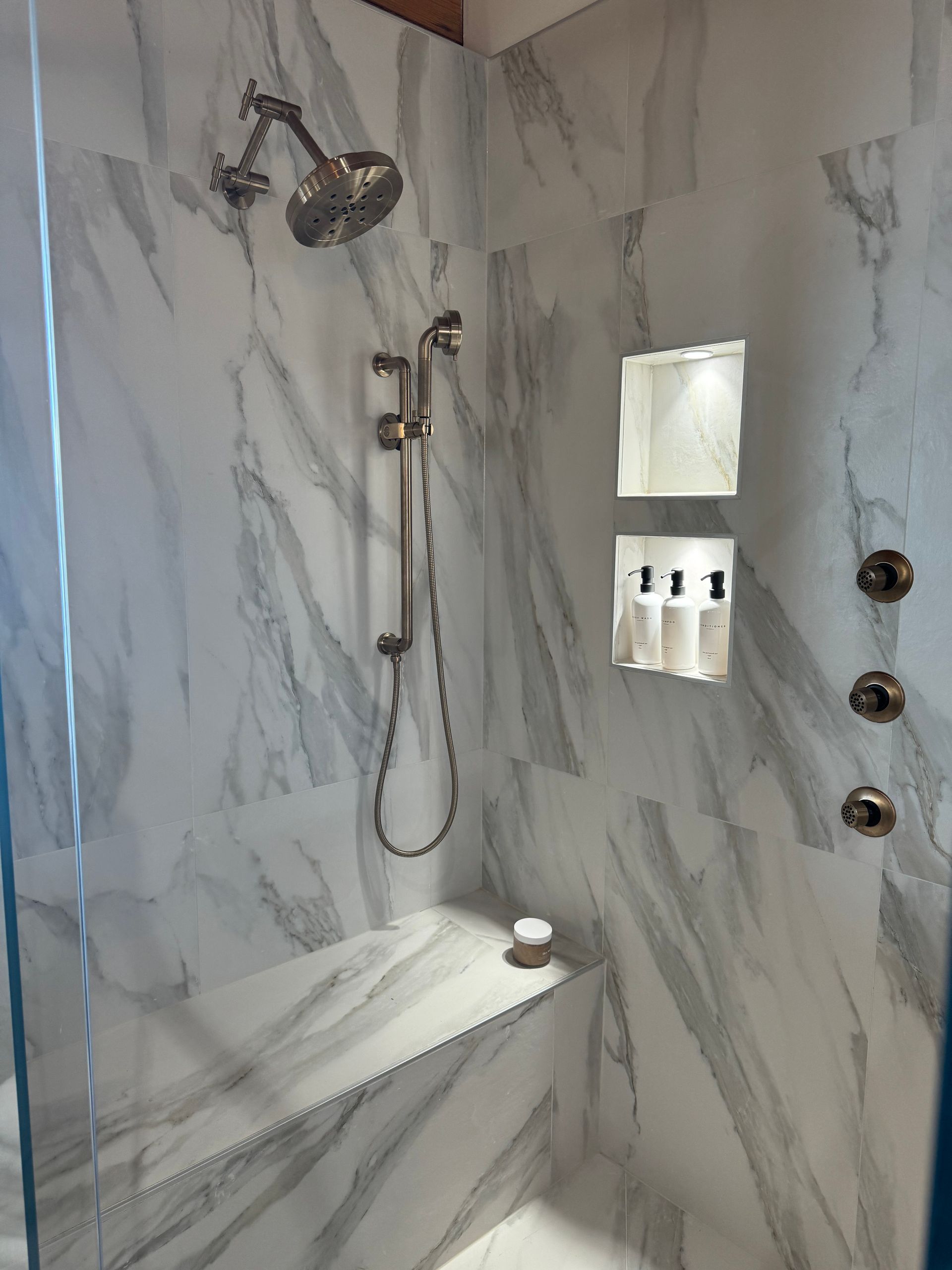 Shower with marble-look tile, fixed showerhead, handheld shower, built-in bench, and shampoo niche with bottles.
