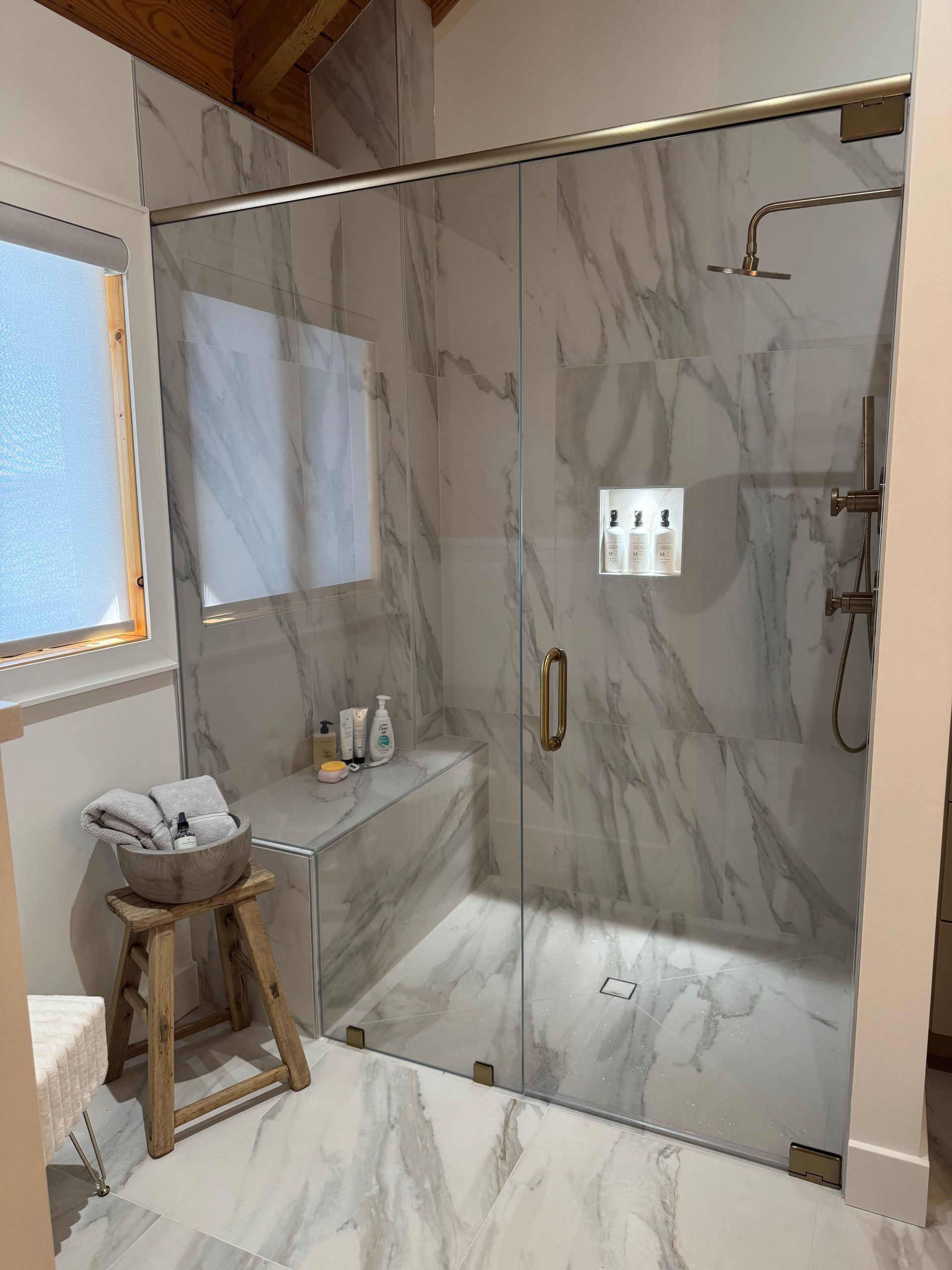 Marble-tiled shower with a glass door, gold fixtures, and a wooden stool with towels, beside a window.