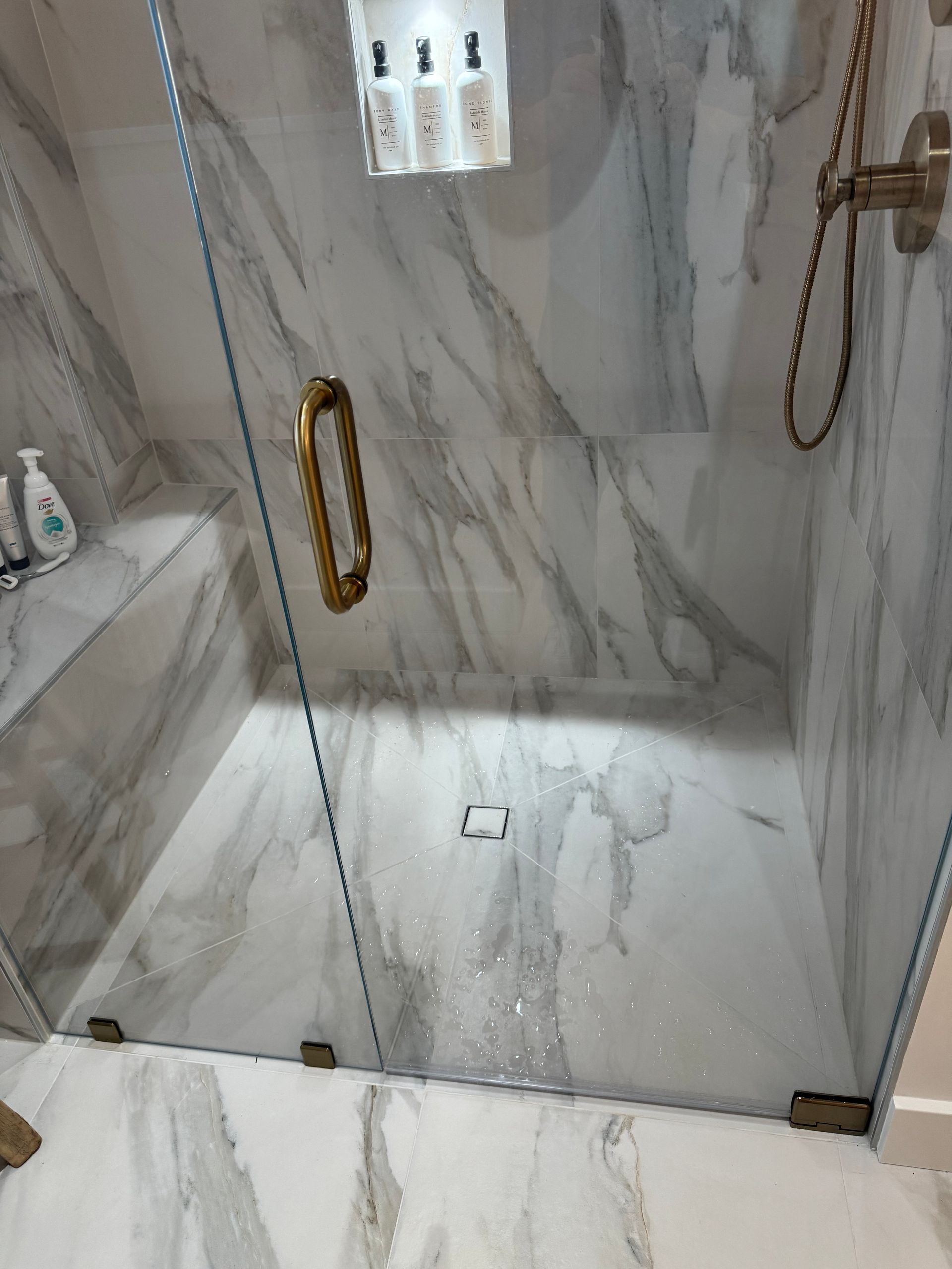 Marble-tiled walk-in shower with glass doors, gold fixtures, and bottles on a shelf.