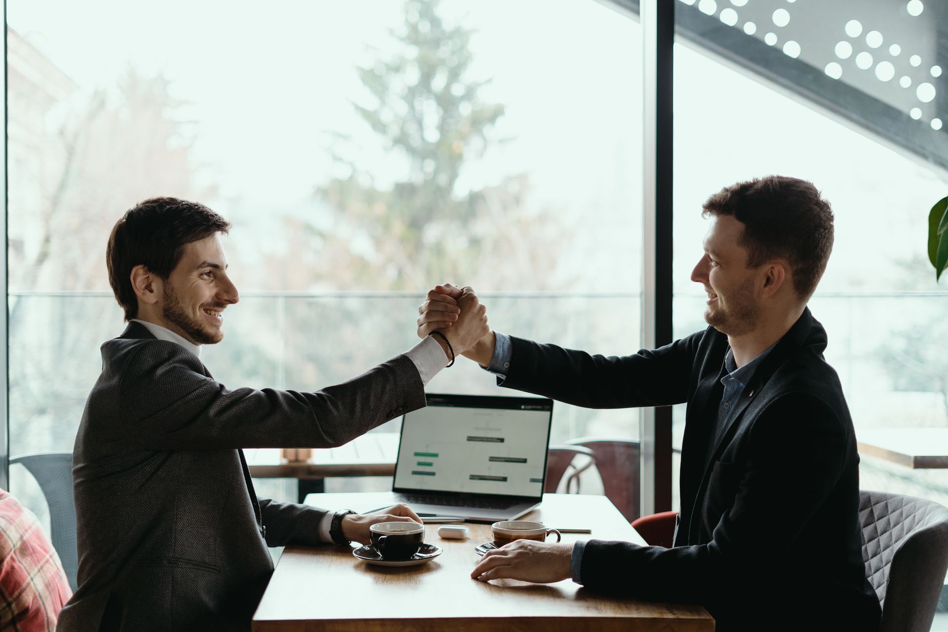 Two men at a table giving a high five, celebrating. Laptop and coffee cup on the table.