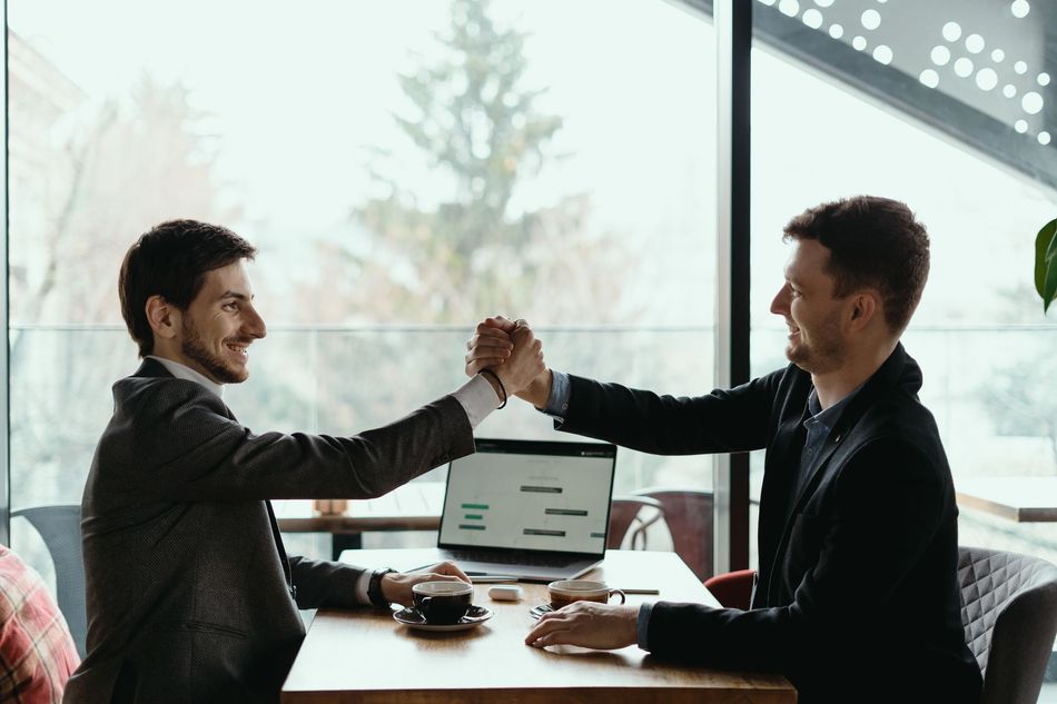 Two men in business attire high-fiving at a table with a laptop and coffee in front of them.