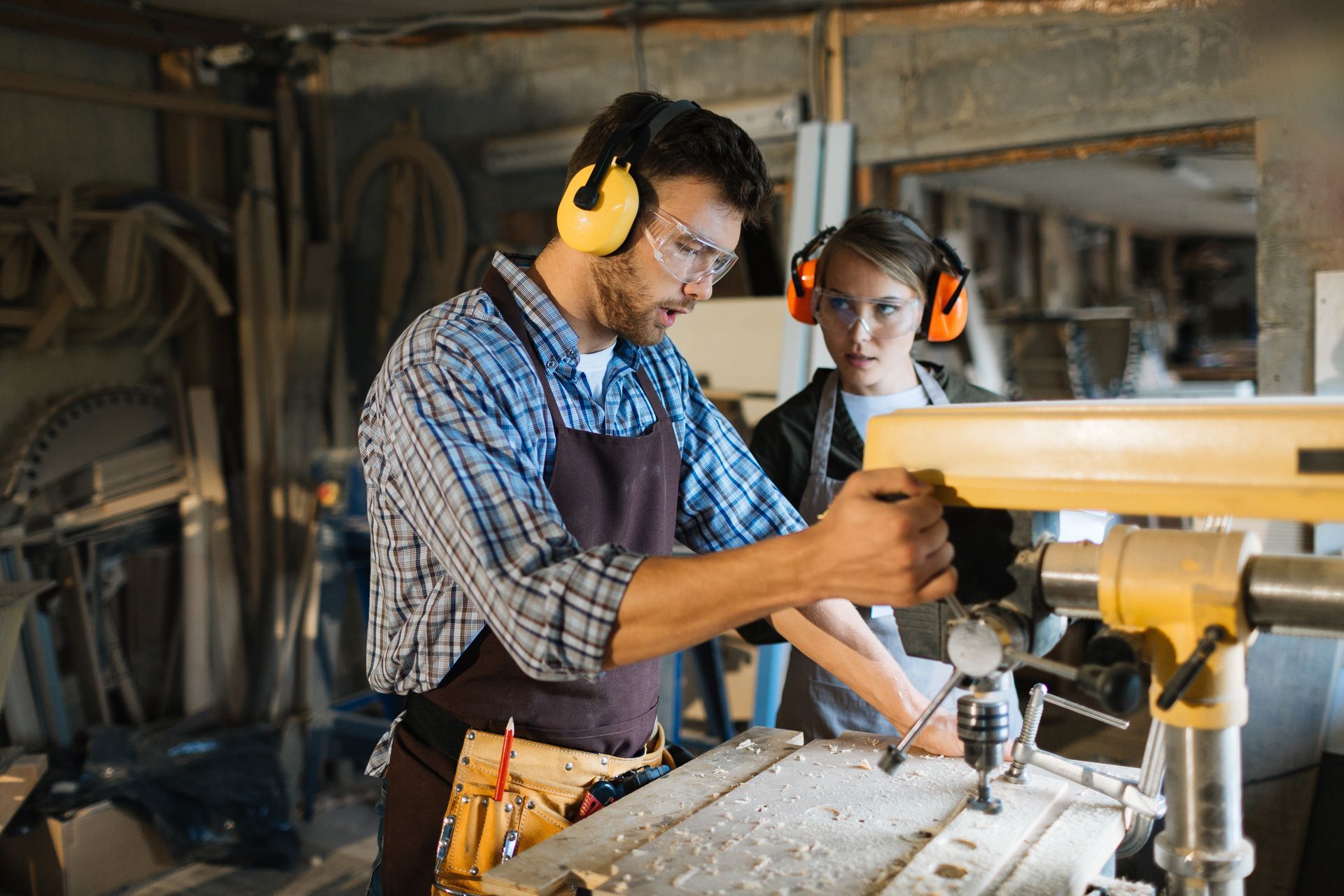 Man and person using a drill press in a workshop, both wearing ear protection.