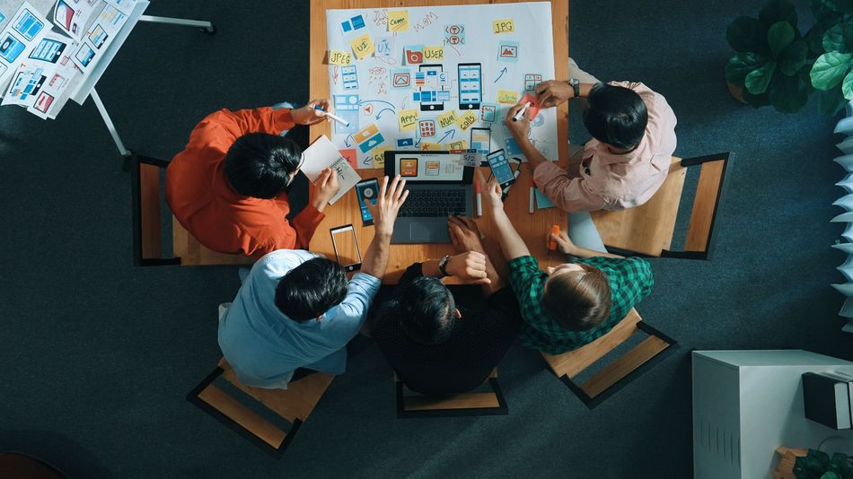 A team of five people collaborating around a table with a laptop and design sketches in an office.