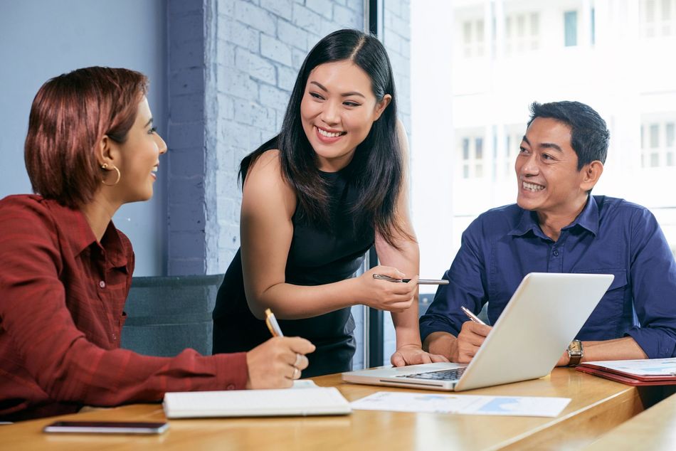 Three diverse colleagues collaborate at a table in an office; smiling and discussing ideas with a laptop and notebooks.