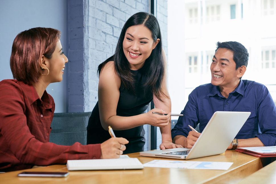 Three diverse colleagues collaborate at a table in an office; smiling and discussing ideas with a laptop and notebooks.