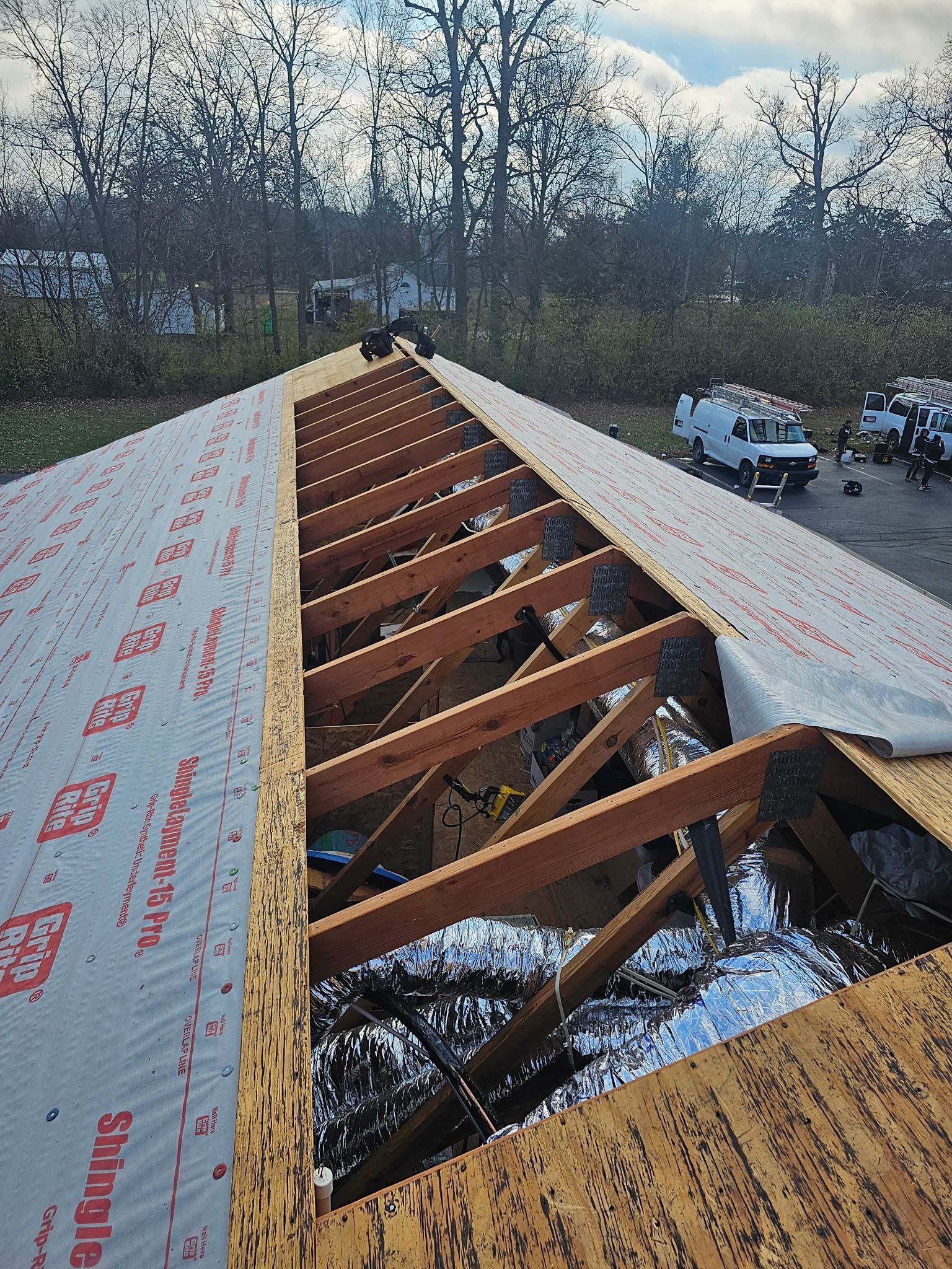 Roof under construction with exposed rafters, insulation, and protective membrane. Trees and vehicles in the background.