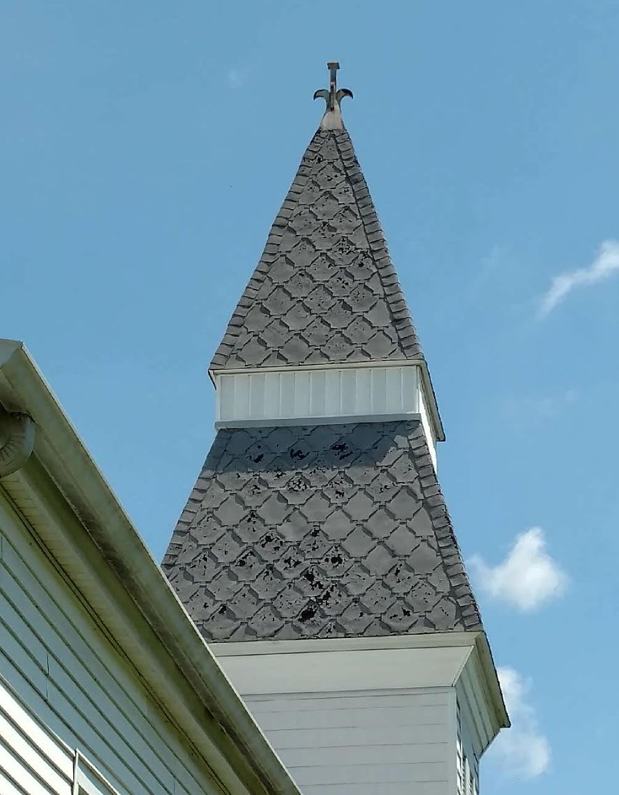 Church steeple with gray shingled roof and cross against a blue sky.