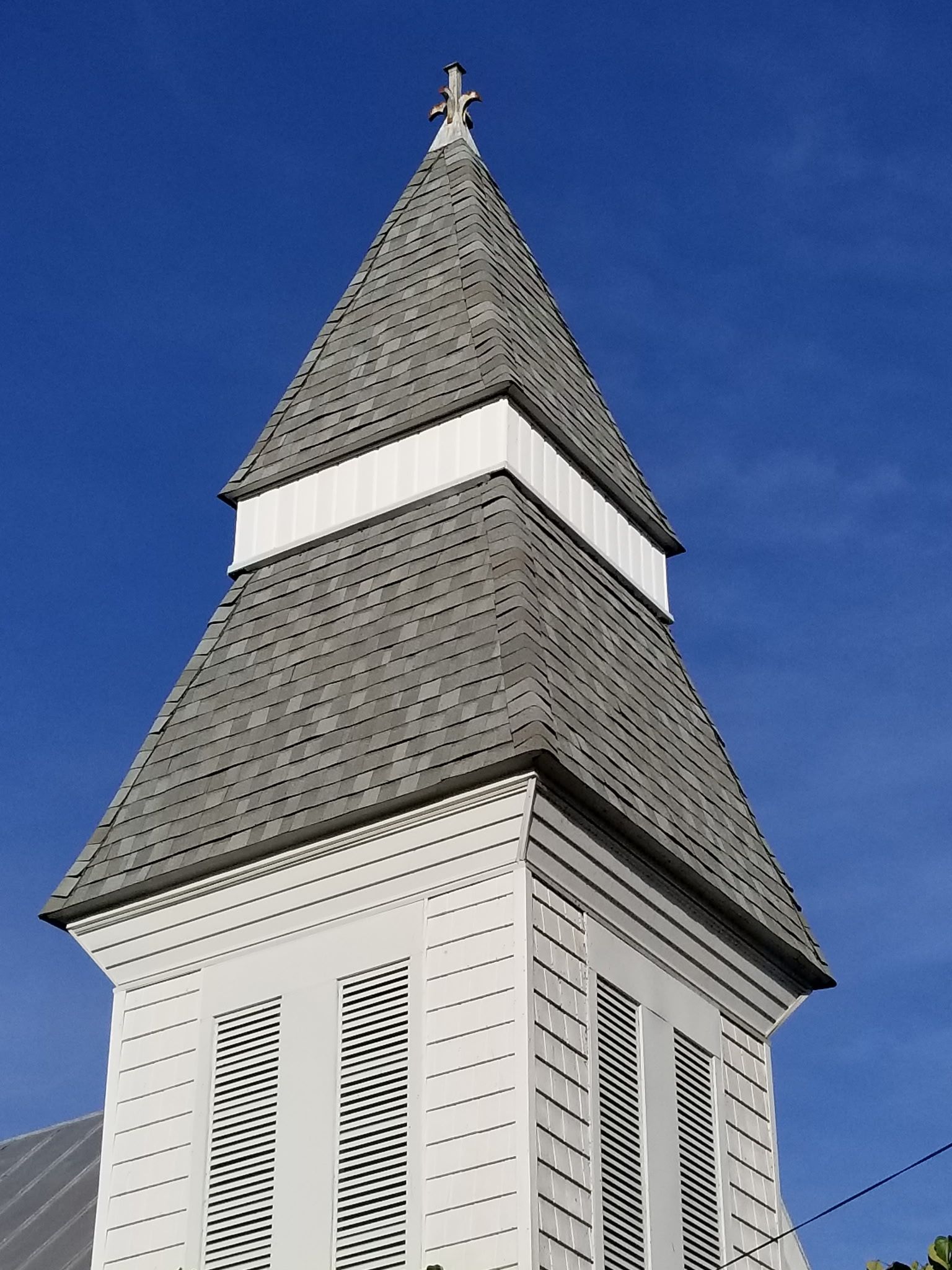White church steeple with gray shingled roof, a cross at the top, against a blue sky.