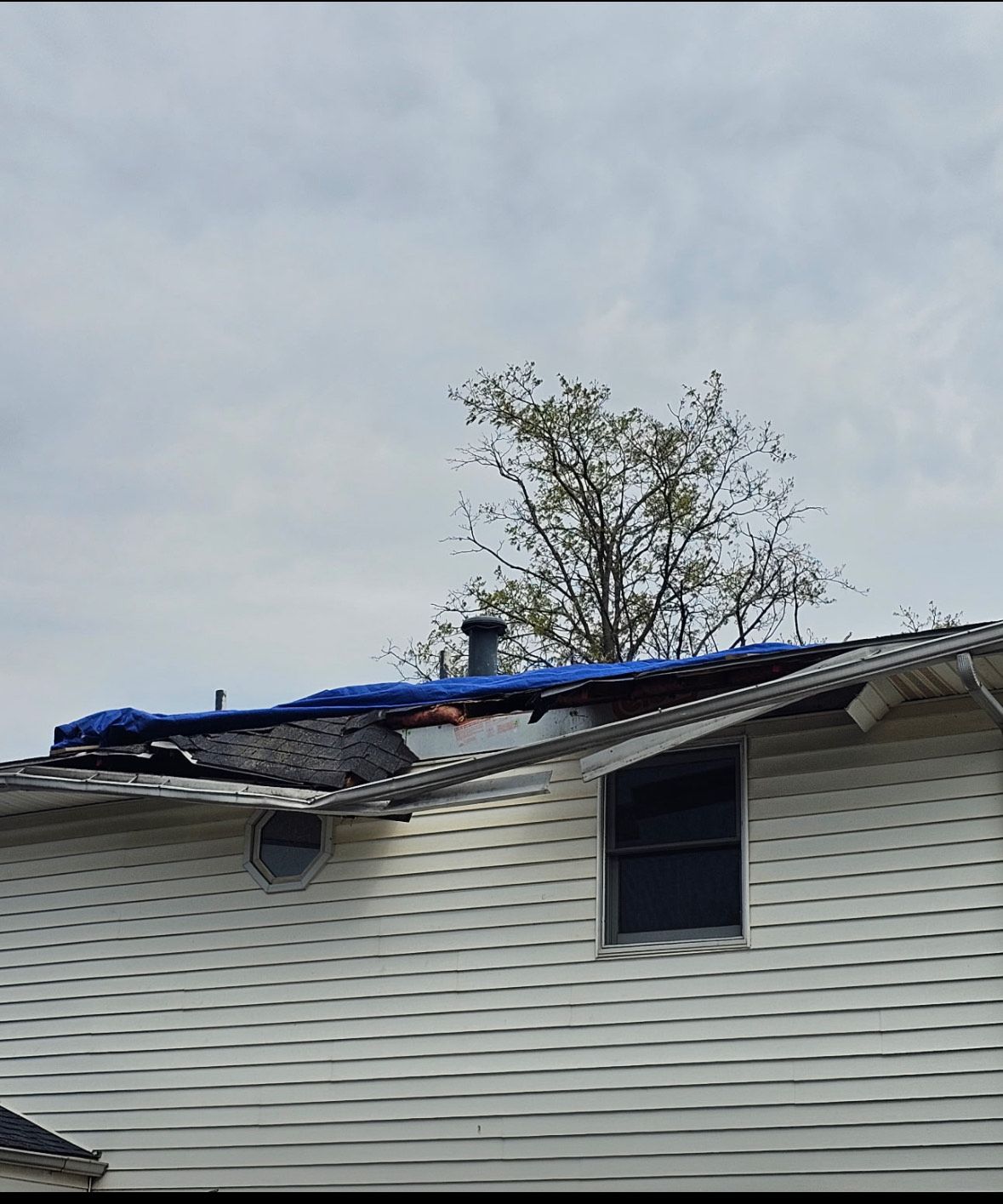House roof with visible damage covered by a blue tarp, with a tree and cloudy sky in the background.