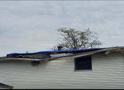 Damaged house roof covered with blue tarp, white siding, cloudy sky, tree in the background.