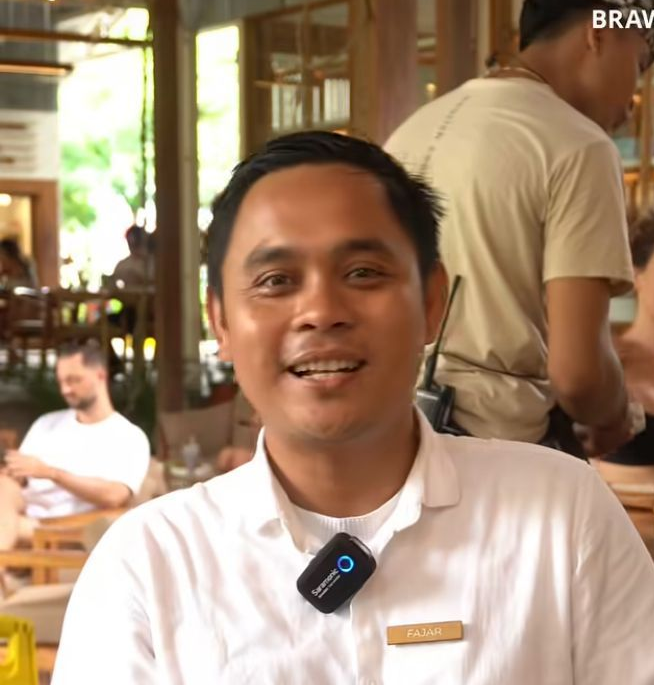 Orang yang tersenyum mengenakan kemeja hitam di sebuah kafe dengan deretan bambu.