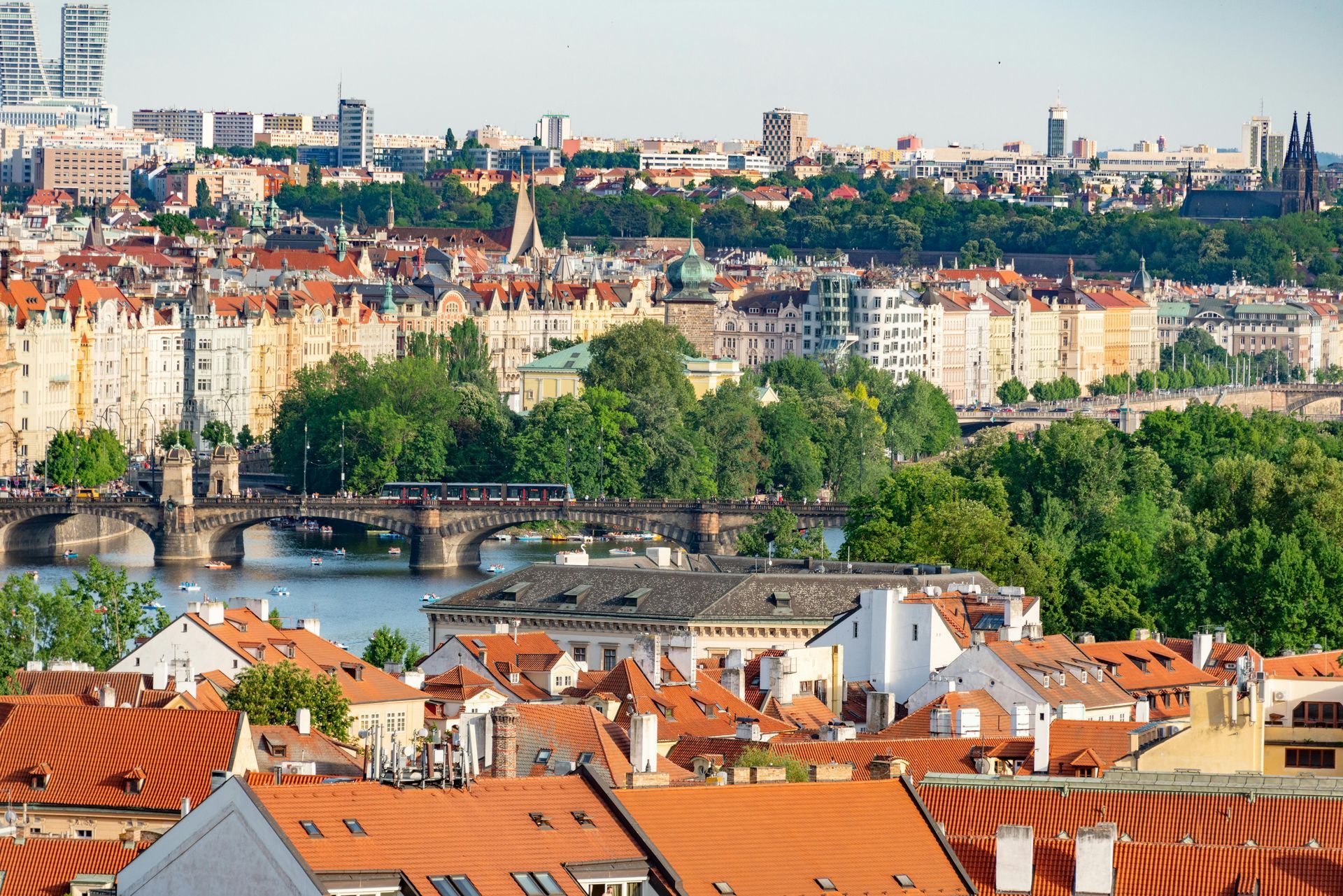 Pemandangan kota Praha dengan atap genteng merah, sungai, jembatan, dan bangunan di bawah langit biru.