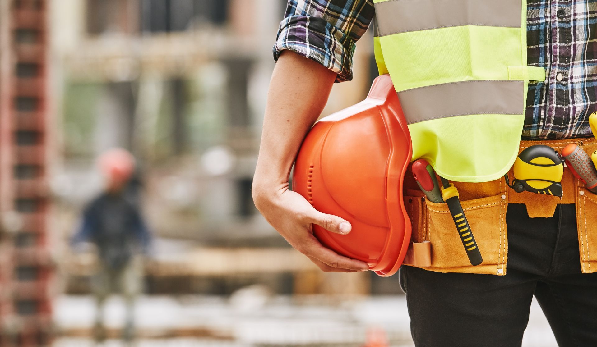 Ouvrier du bâtiment sur un chantier, tenant un casque orange et portant un gilet de sécurité.