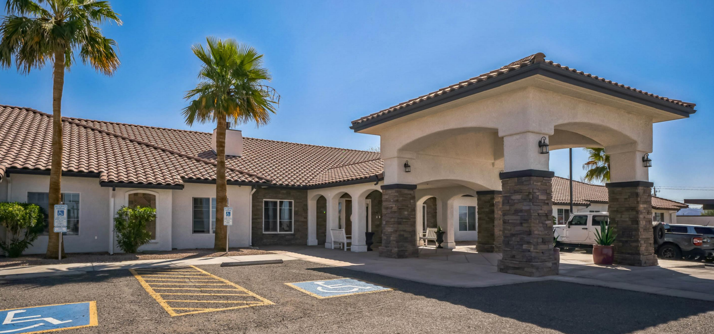 A beige, single-story building with a covered entrance; palm trees in front under a blue sky.